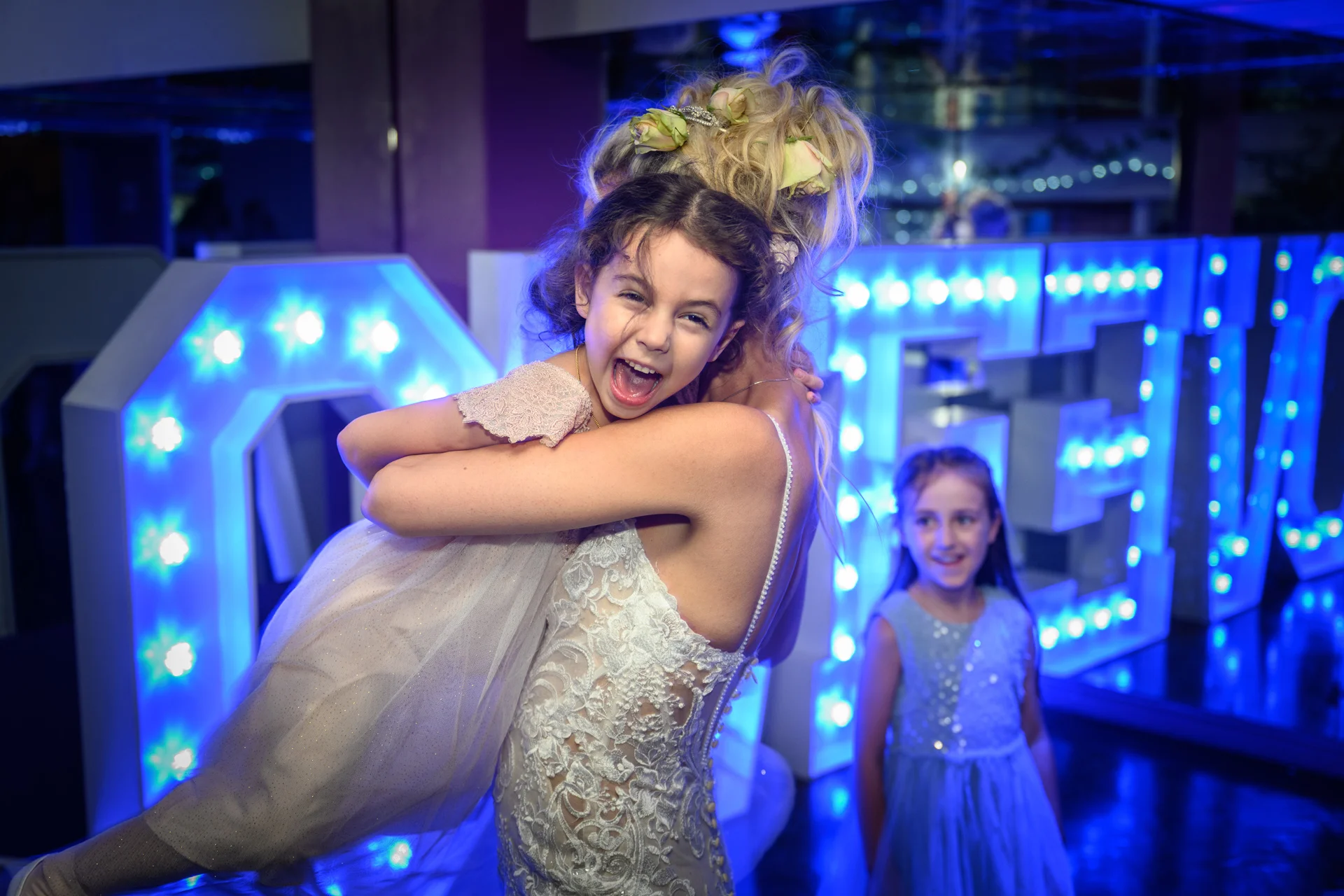 A joyful bride lifts a laughing flower girl into the air in front of glowing blue light-up letters during the evening reception.