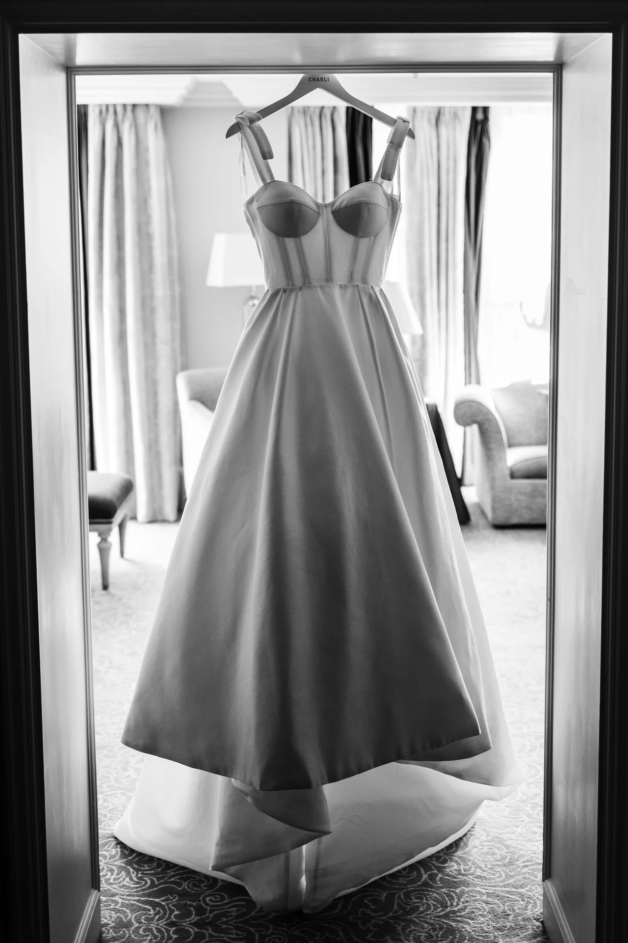 A structured satin wedding dress with a corset bodice hangs on a personalised hanger within a doorway during morning bridal preparations.