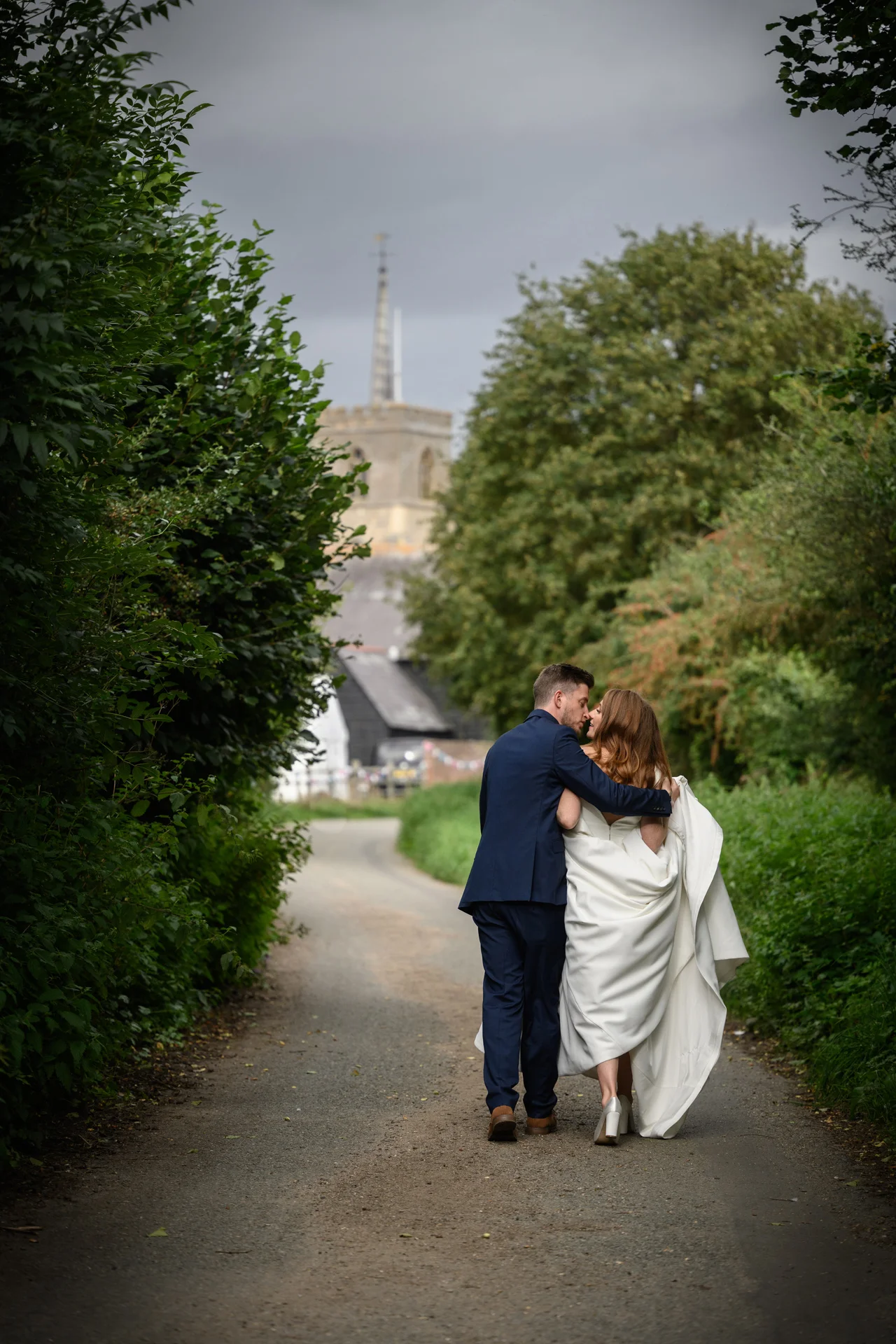 A newlywed couple walks hand in hand down a rural lane lined with green trees and hedges, with a traditional church tower visible in the distance under an overcast sky.