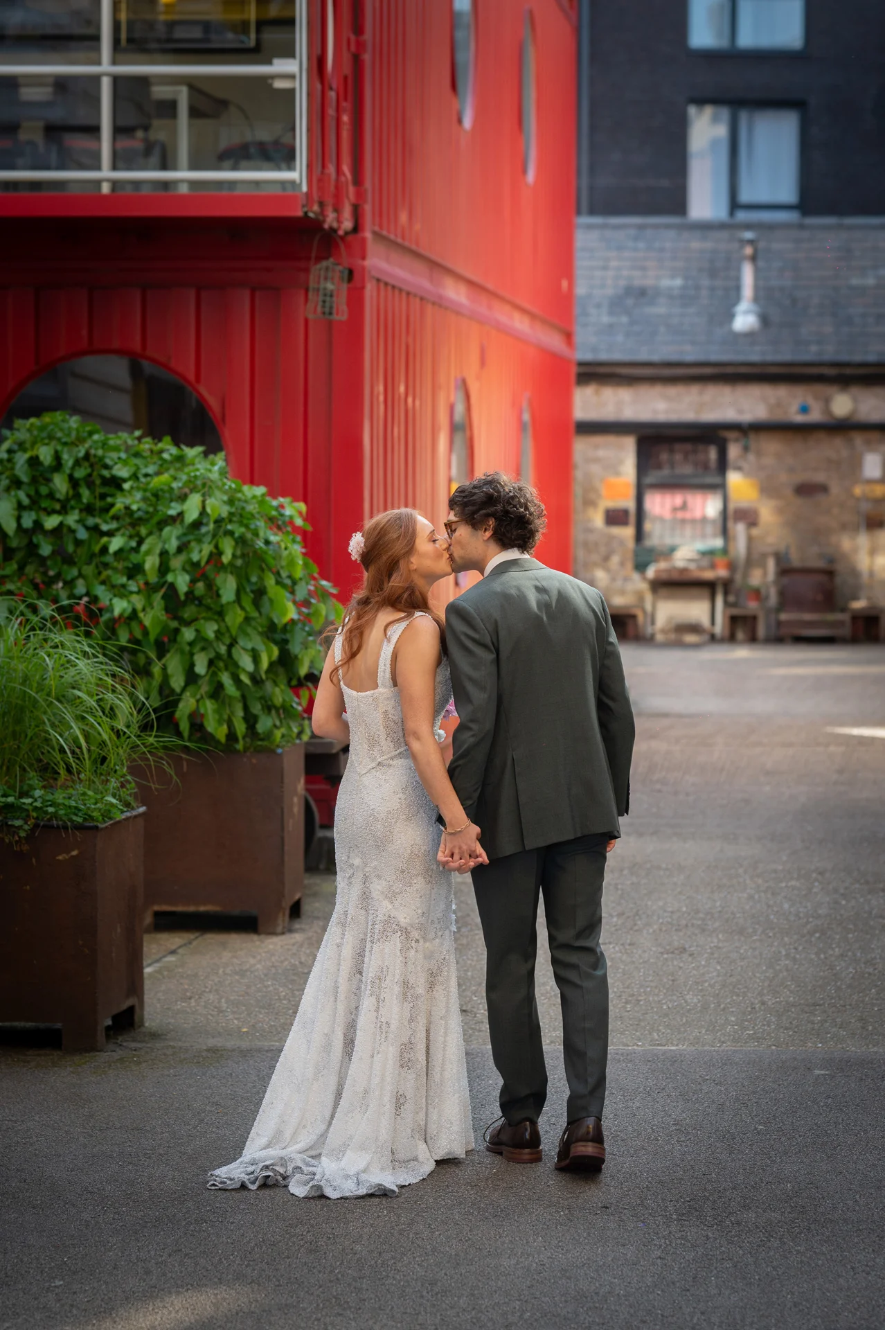 A bride in a lace fit-and-flare gown and a groom in a green suit share a tender kiss while holding hands in an industrial courtyard next to a striking red building.