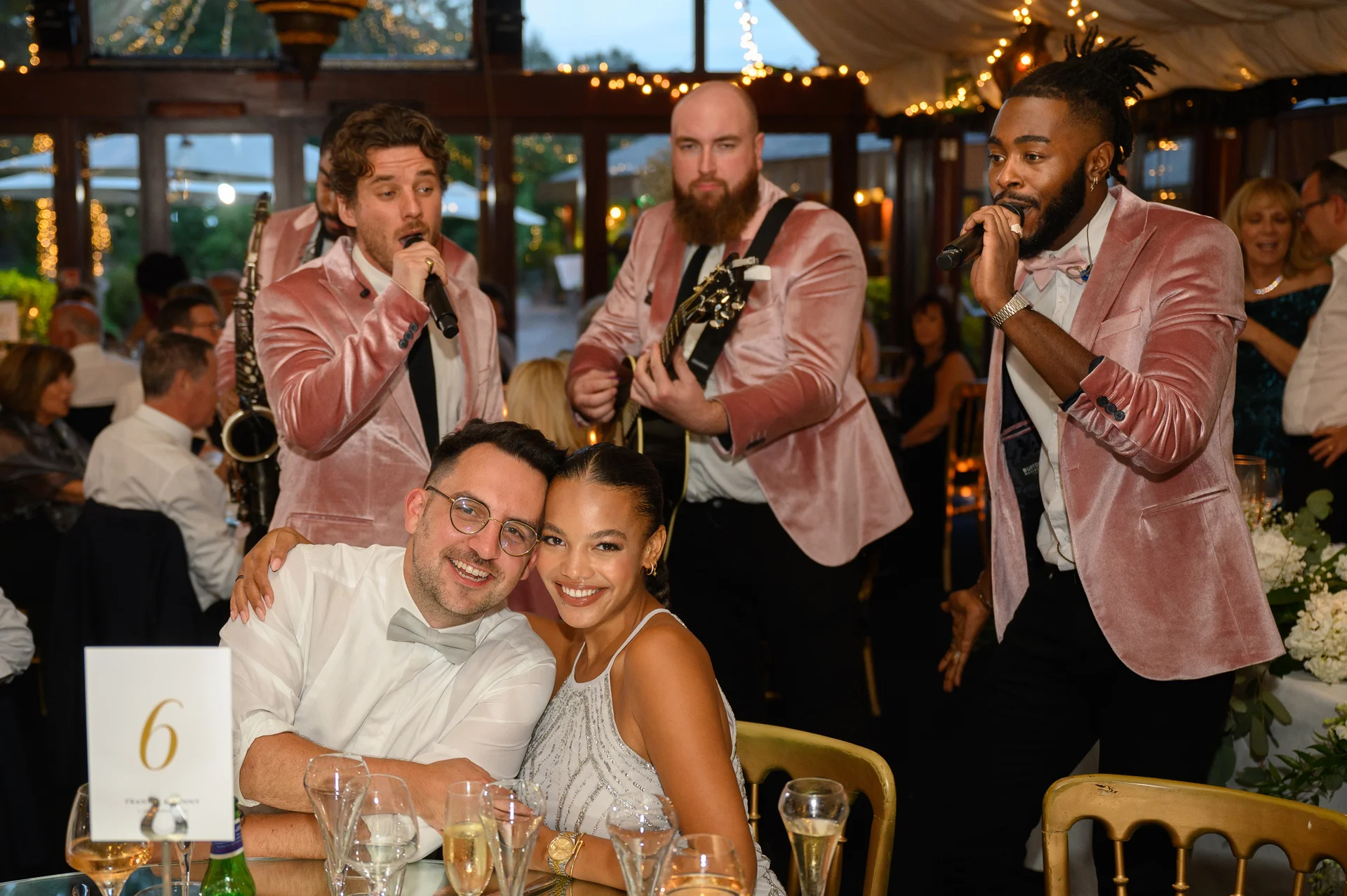 A smiling bride and groom embrace at their wedding reception while a live band wearing pink velvet jackets performs nearby.