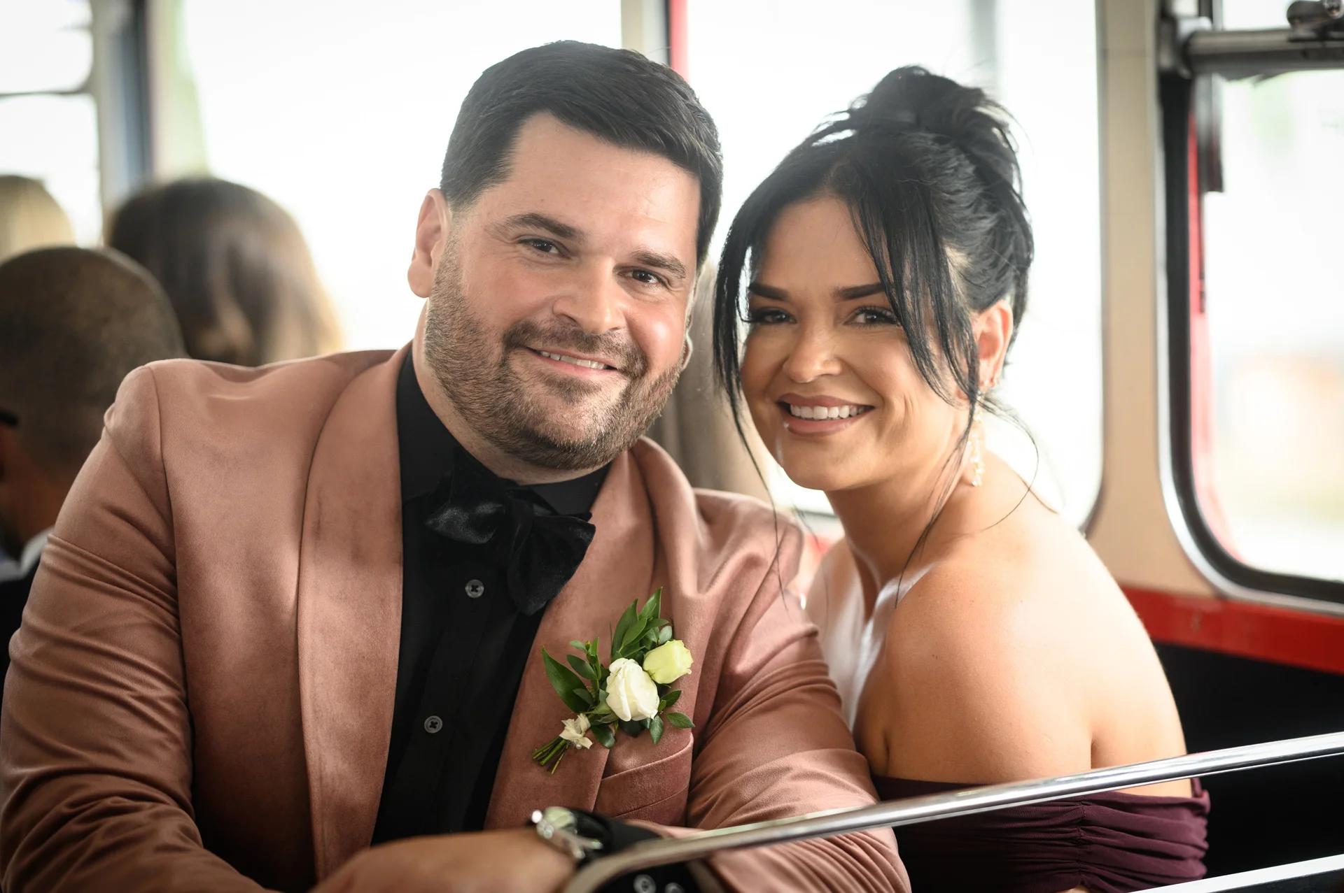 A smiling bride and groom pose together inside a classic red double decker bus, with the groom wearing a velvet dinner jacket and a floral buttonhole.