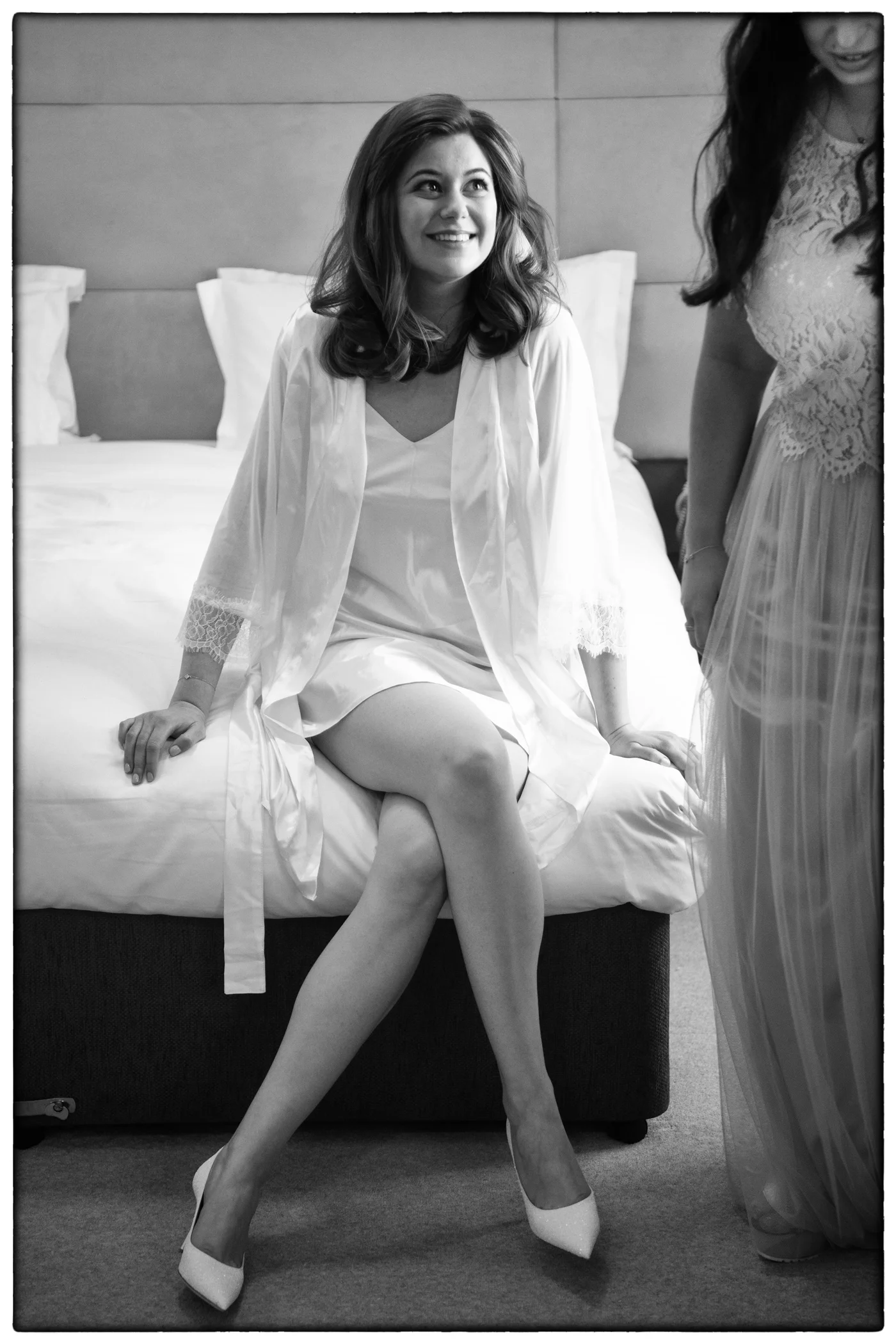 A smiling bride in a white robe and heels sits on a bed while getting ready, accompanied by someone standing nearby in a lace dress.