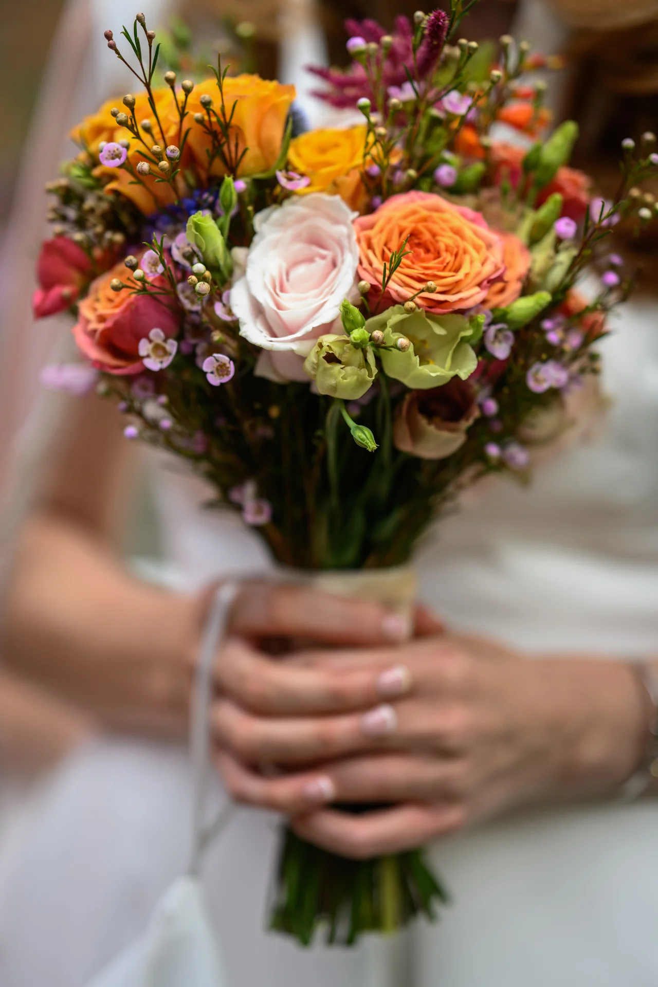 A close-up shot of a bride holding a vibrant, hand-tied bouquet featuring orange roses, a soft pink center rose, and delicate wildflowers.