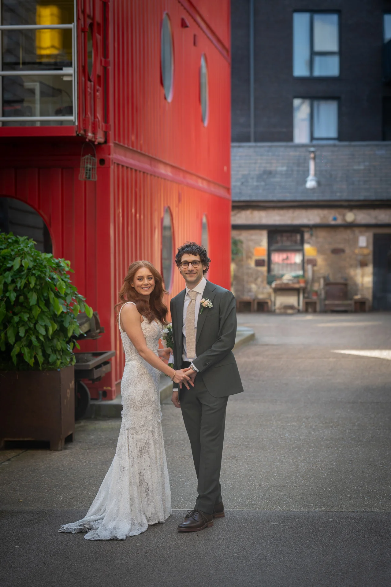 A bride in a lace sheath dress and a groom in a dark grey suit stand hand in hand outdoors in a paved courtyard against a striking red container building.