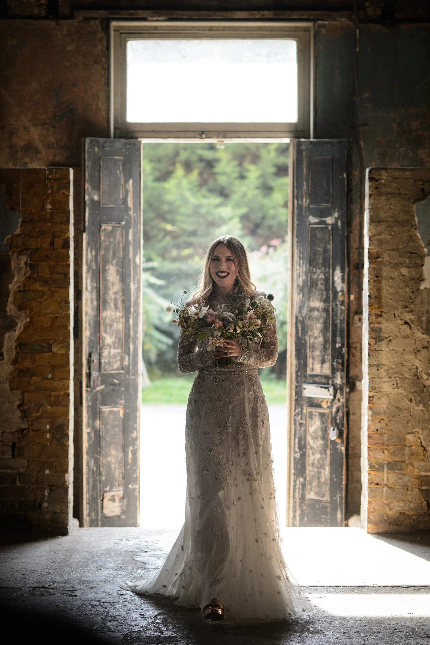 A smiling bride stands framed in large rustic open doors, holding a textured wildflower bouquet while wearing a beautifully embellished long-sleeve gown inside an industrial venue.