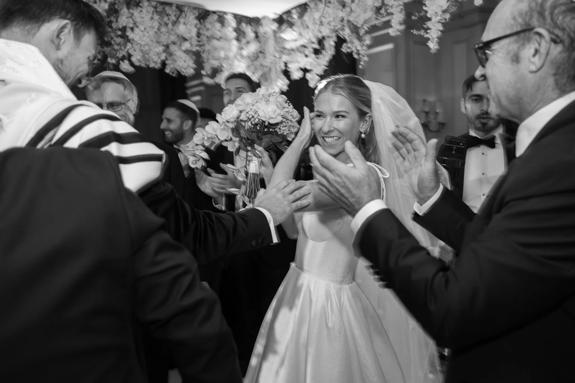 A joyful bride laughs and holds her bouquet as she is congratulated by applauding guests during a celebratory wedding moment.