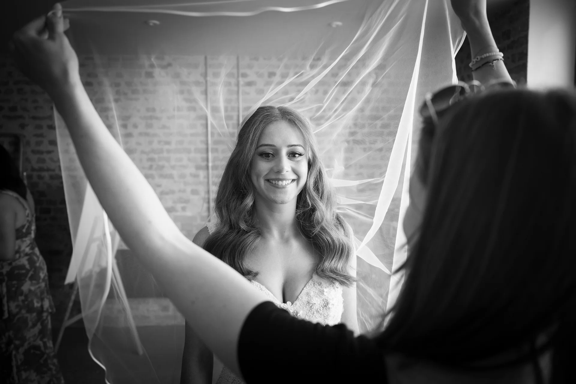 A smiling bride looks through a sheer veil held up by people during morning preparations in a room with exposed brick walls.