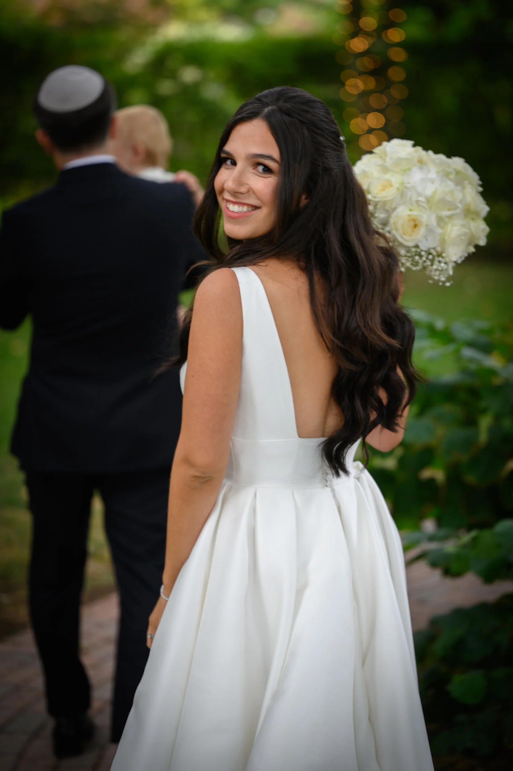A smiling brunette bride in a white satin gown with a low back looks back over her shoulder while holding a white rose bouquet outdoors.