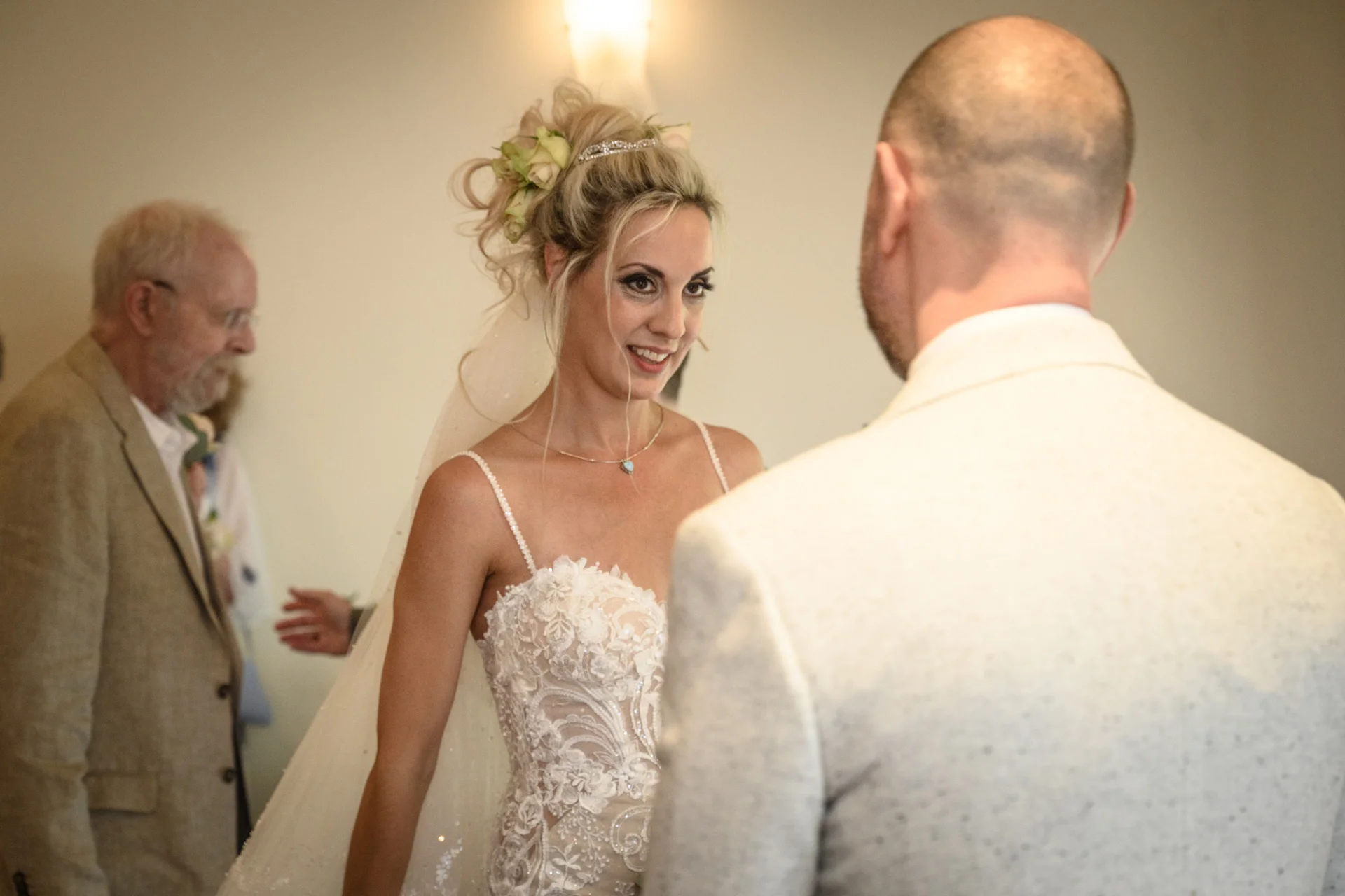 A smiling bride in a detailed lace gown and veil looks lovingly at her groom during the wedding ceremony, while a guest looks on from the background.