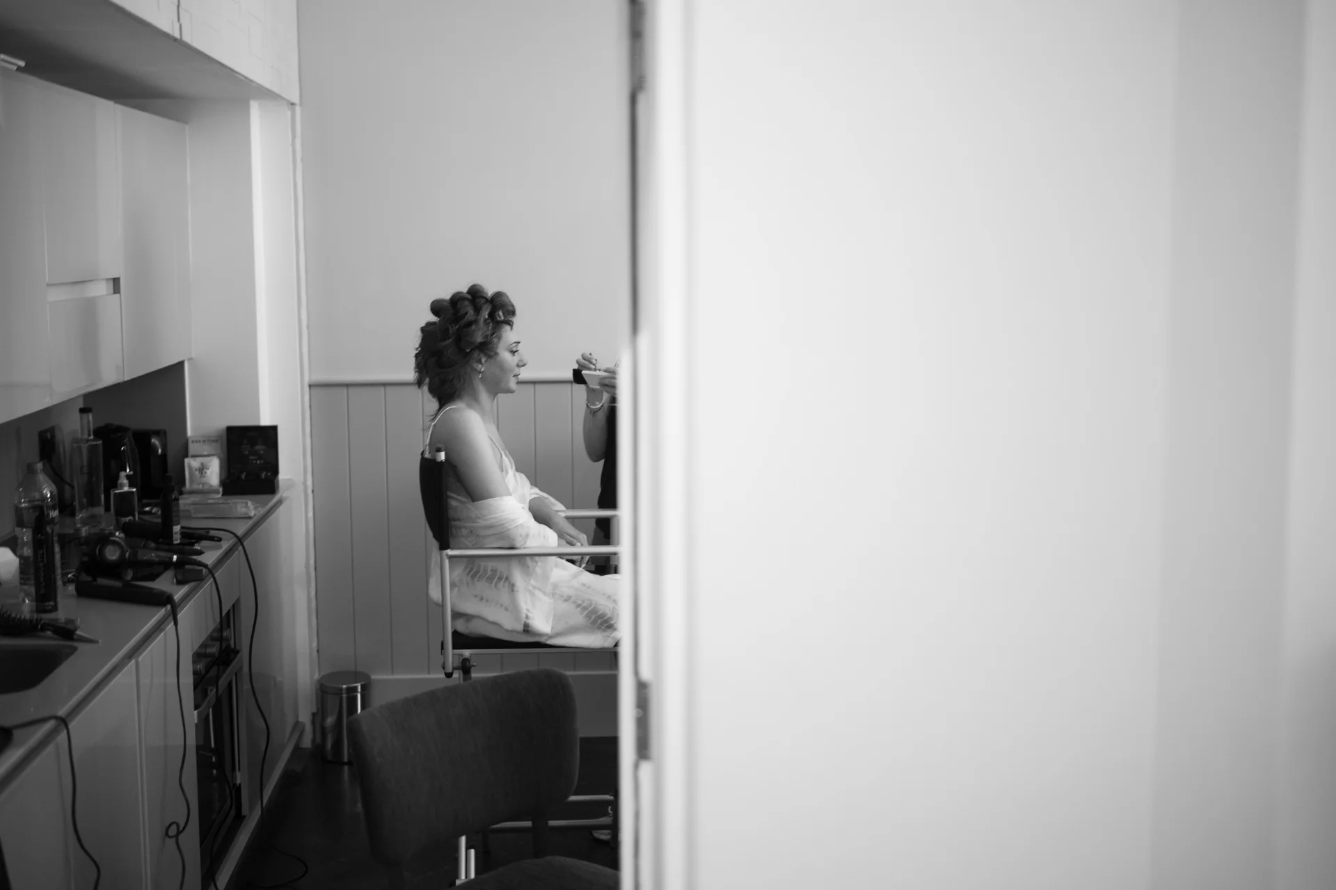 A bride sits in a styling chair wearing a robe with hair rollers while getting her makeup applied, viewed through a doorway in black and white.