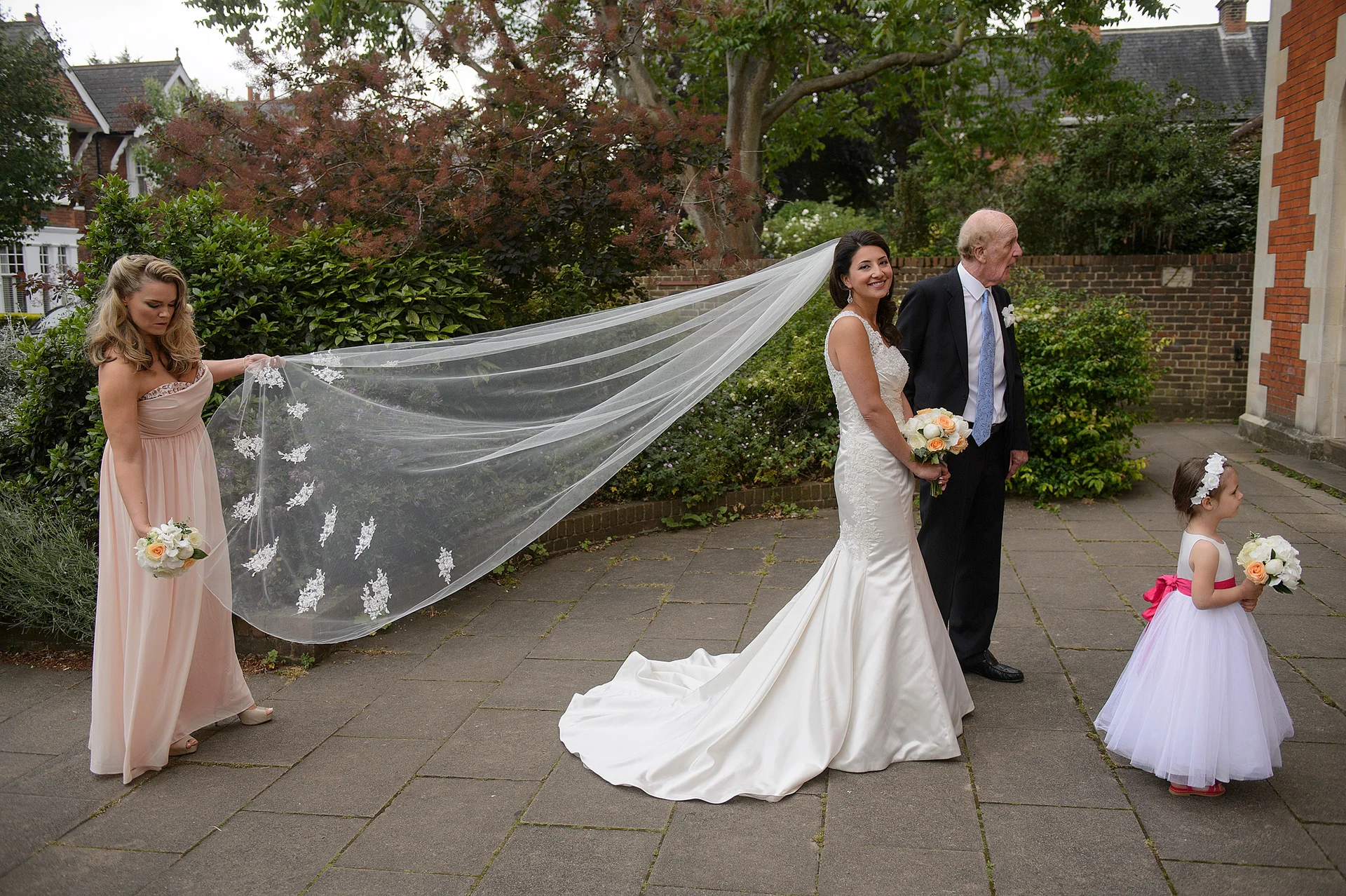 A smiling bride in a satin fit-and-flare gown stands alongside her father outside a church while a bridesmaid holds her long lace-edged veil.