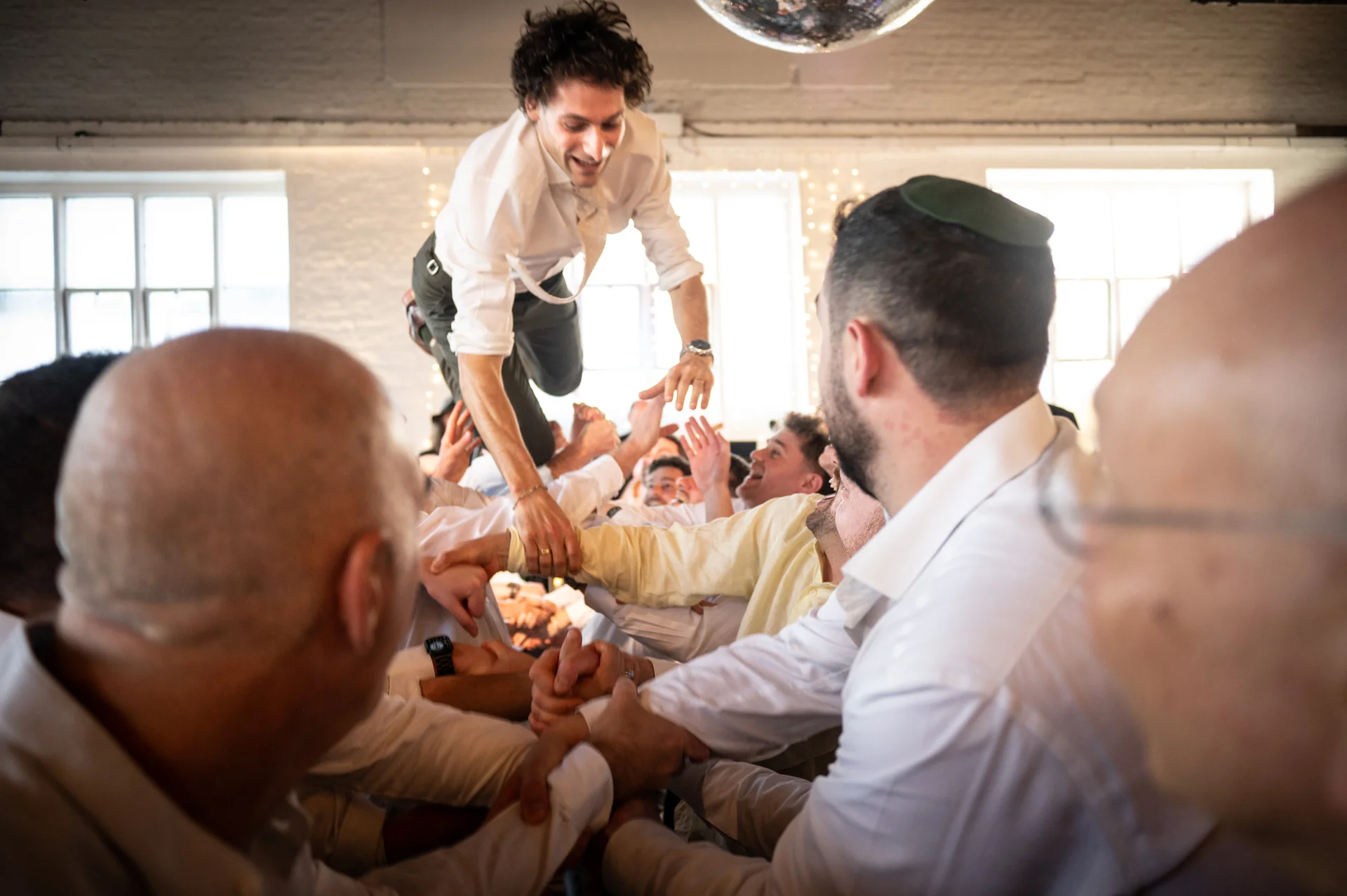 A groom is lifted high into the air by cheering wedding guests during a joyful celebration underneath a glittering disco ball.