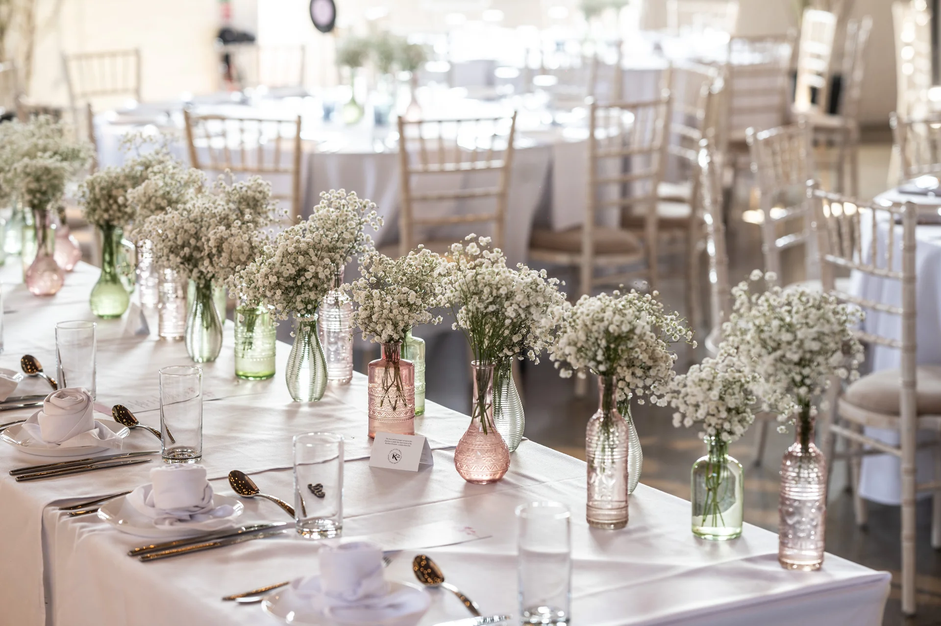 A long banquet table set for a wedding breakfast, beautifully decorated with rows of delicate baby's breath flowers in pink and green bud vases.