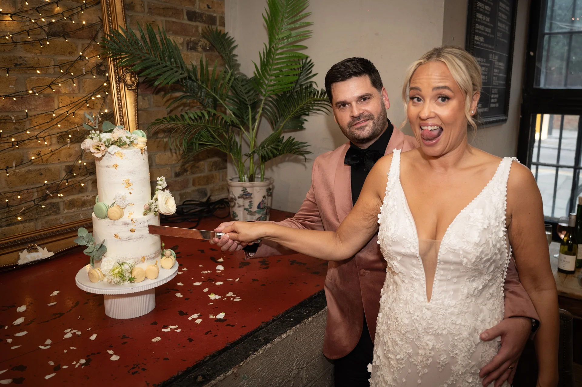 A newly married couple laugh playfully while cutting their two-tier semi-naked wedding cake decorated with gold leaf and macarons against an exposed brick backdrop.
