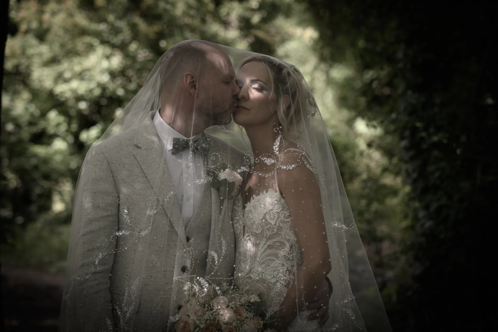 A romantic couple shares a tender kiss beneath an intricately embroidered veil while standing together in a lush woodland setting.