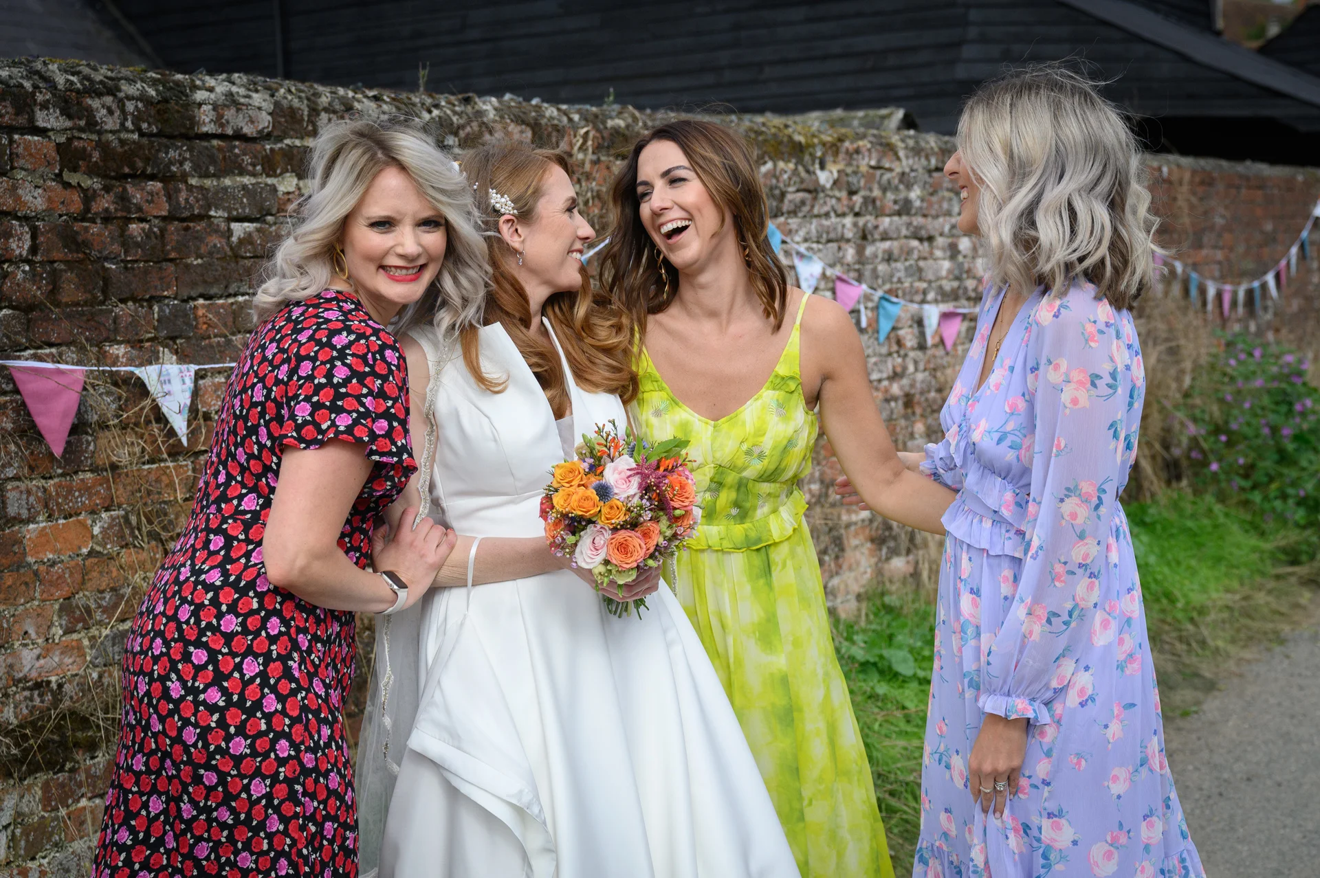 A joyful bride holding a colourful bouquet shares a hearty laugh with her three bridesmaids dressed in vibrant floral dresses outdoors against an old brick wall.