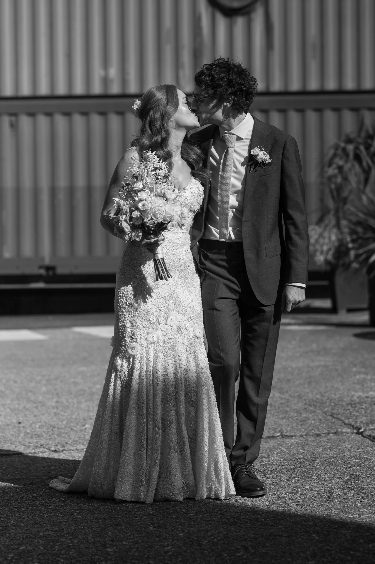 A newlywed couple shares a tender kiss in bright sunlight outside an industrial venue, with the bride holding a textured bouquet.