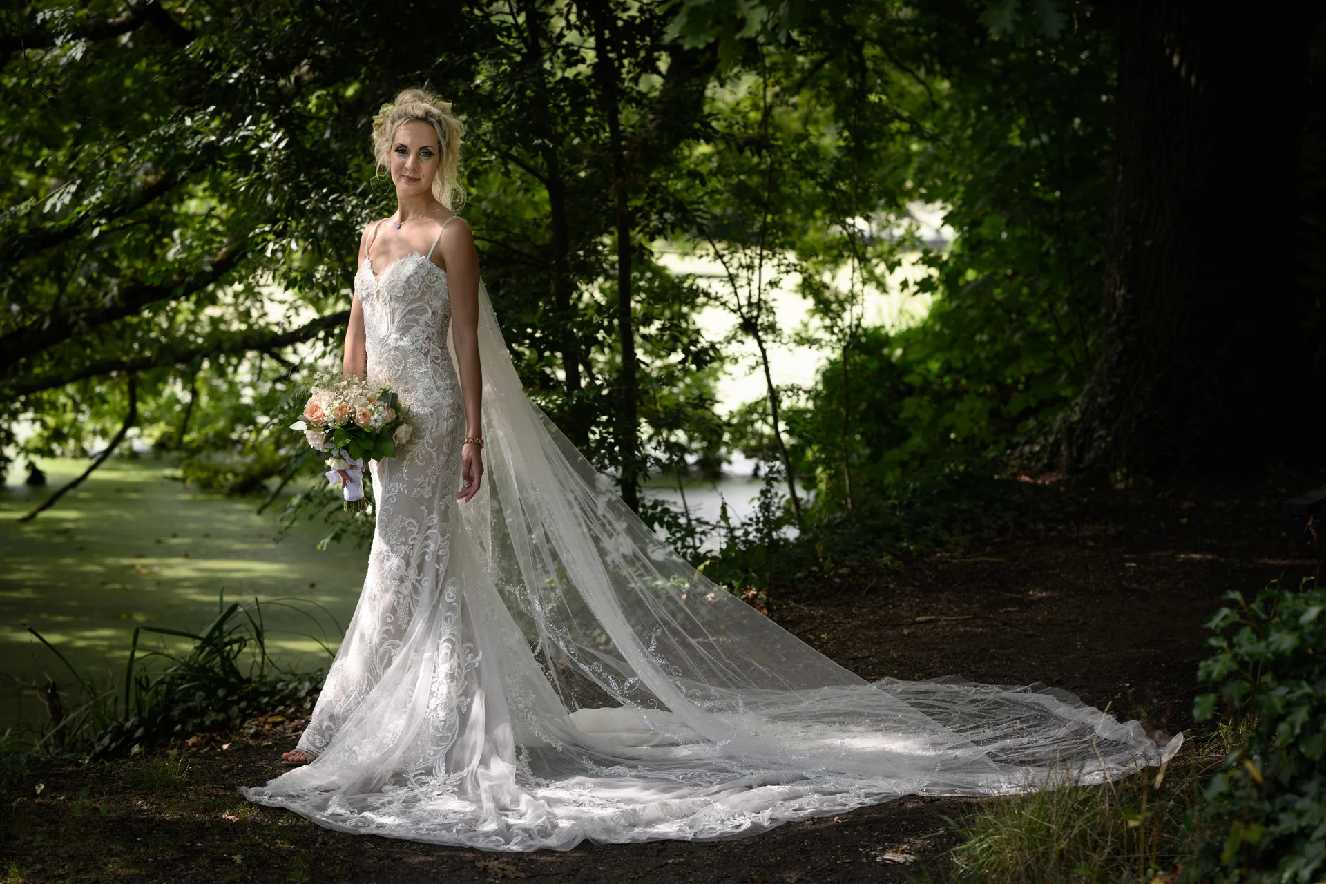 A stunning bride stands along a woodland path beside a lake, wearing a fitted lace gown with a dramatic cathedral veil and holding a delicate bouquet.