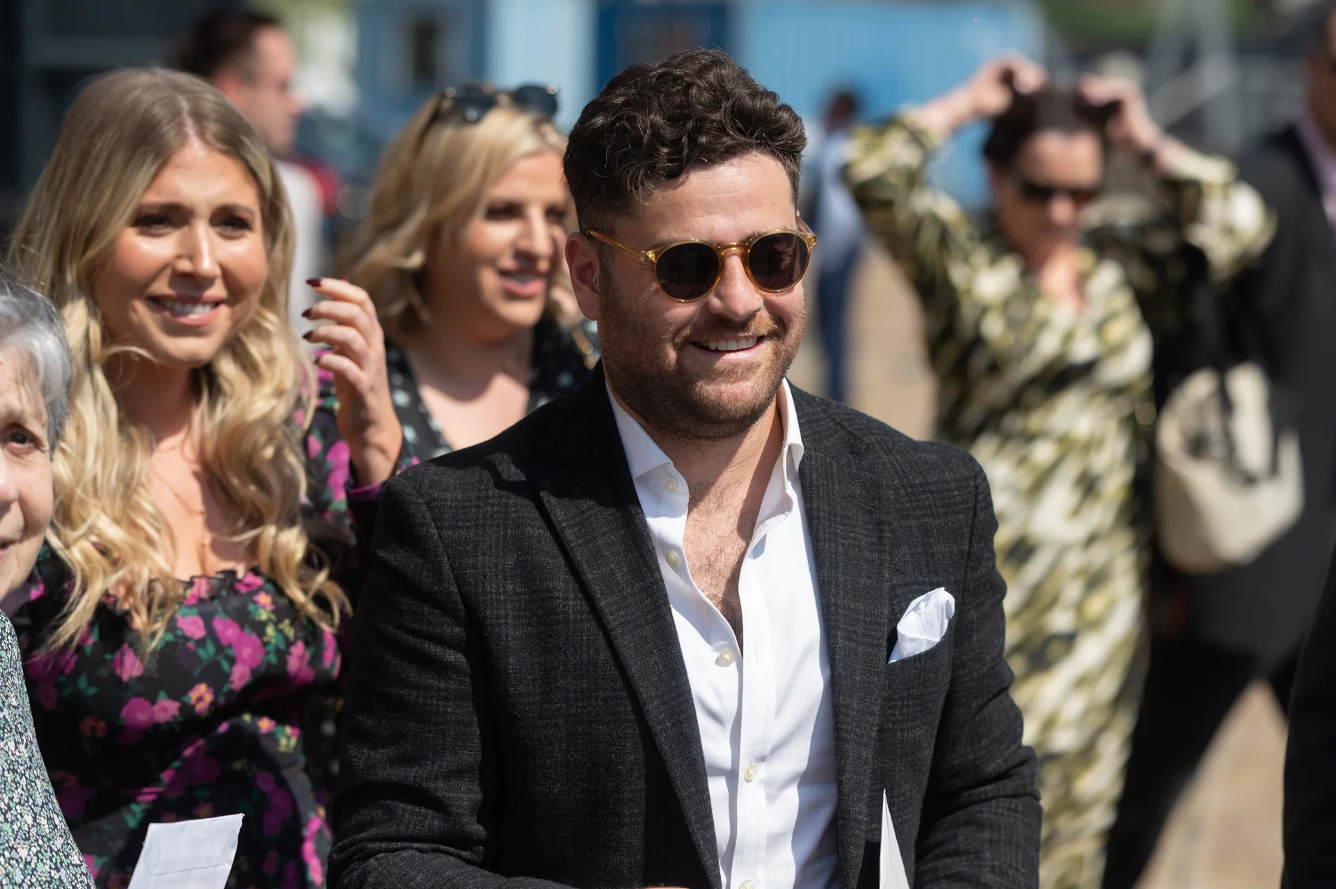 A stylishly dressed male guest wearing a charcoal suit, white shirt, and sunglasses smiles candidly while mingling outdoors surrounded by wedding guests in the sunshine.