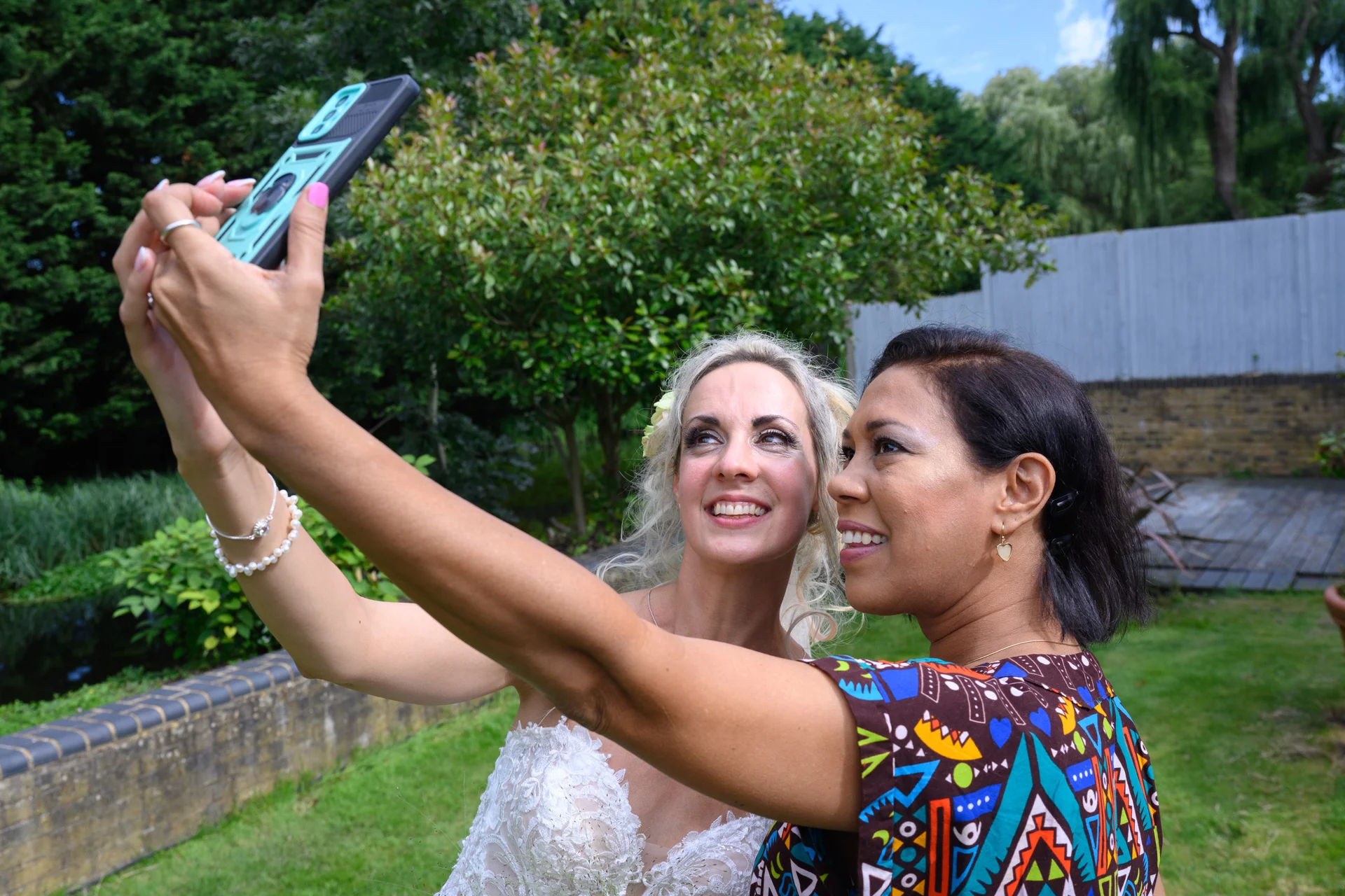 A bride in a lace wedding dress and a guest take a cheerful selfie together outdoors in a lush garden setting.
