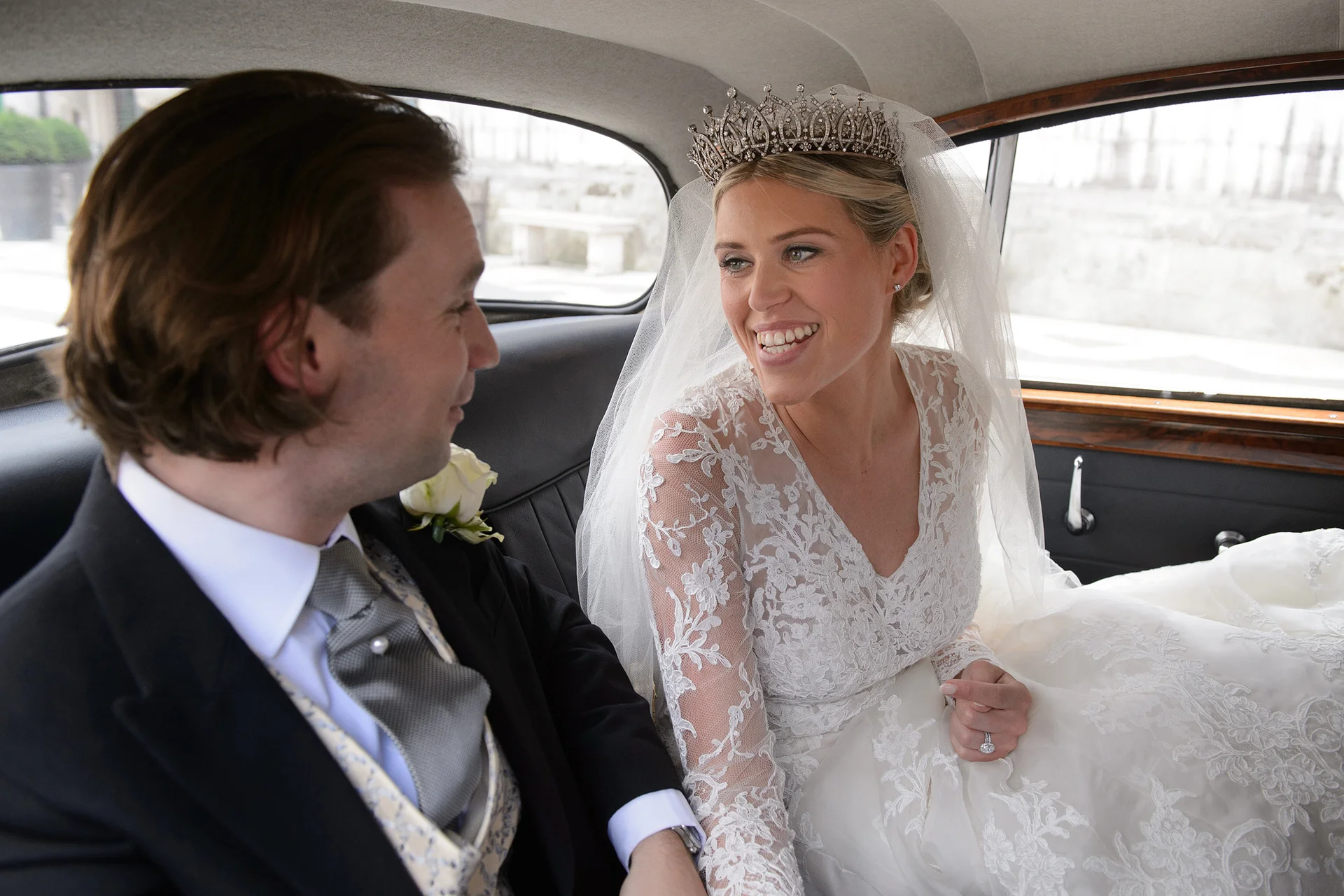 A newlywed couple shares a joyful, smiling glance while seated in the back of a classic wedding car.