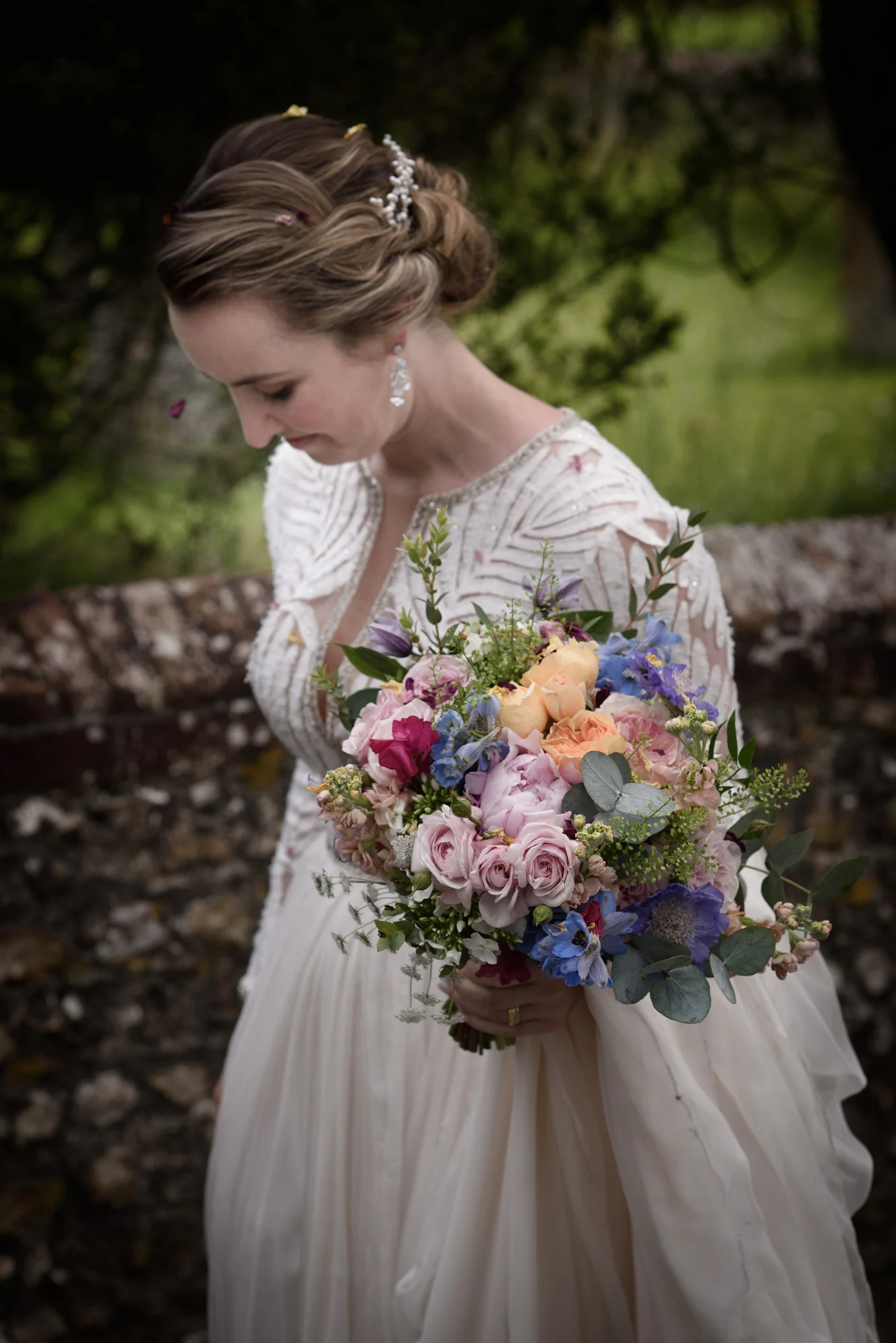 A stunning bride with an intricate updo looks down at her colourful, hand-tied garden bouquet, wearing an embroidered long-sleeve gown against a rustic stone wall.