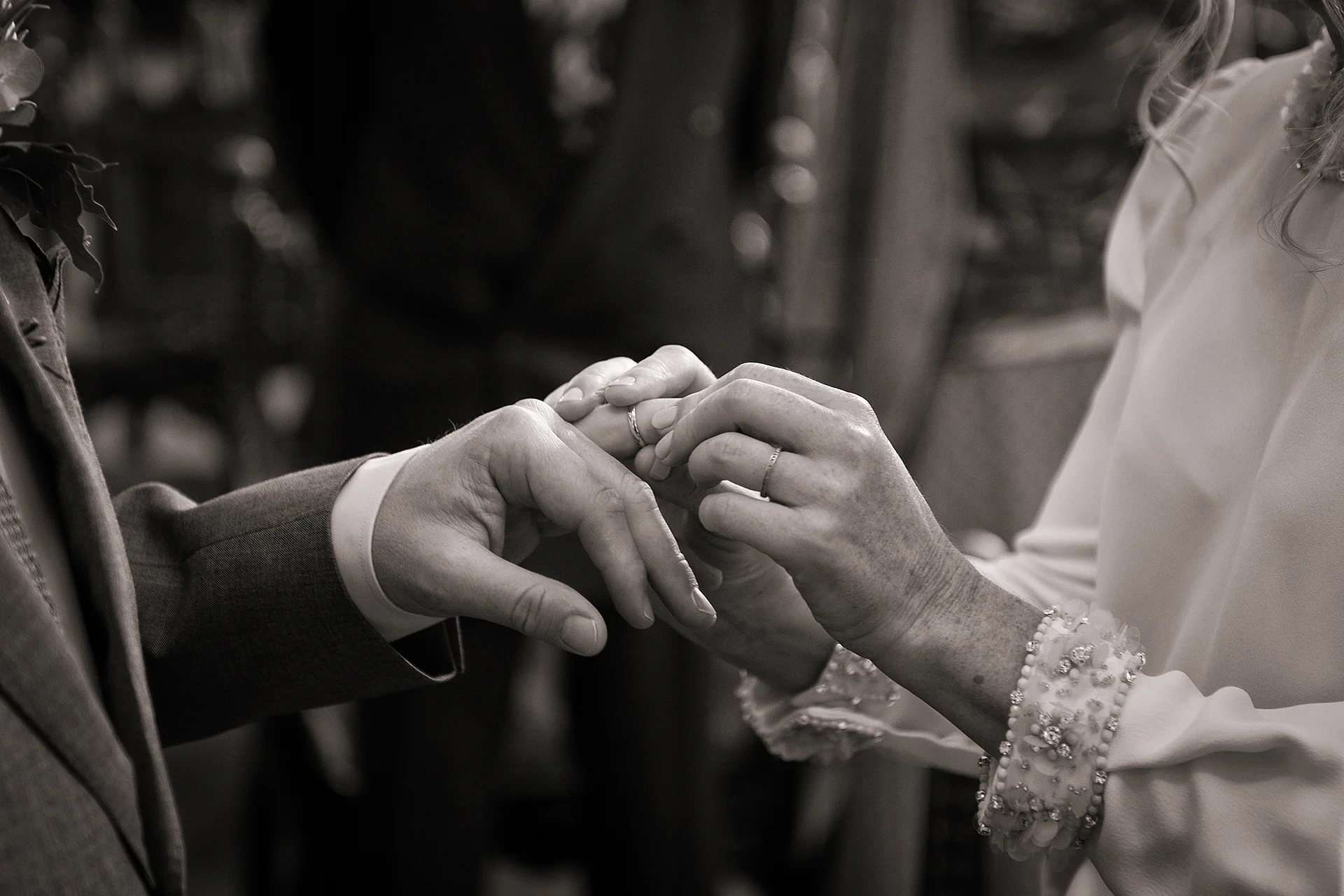 A close-up black and white shot captures the tender moment of the couple exchanging wedding rings during the ceremony.