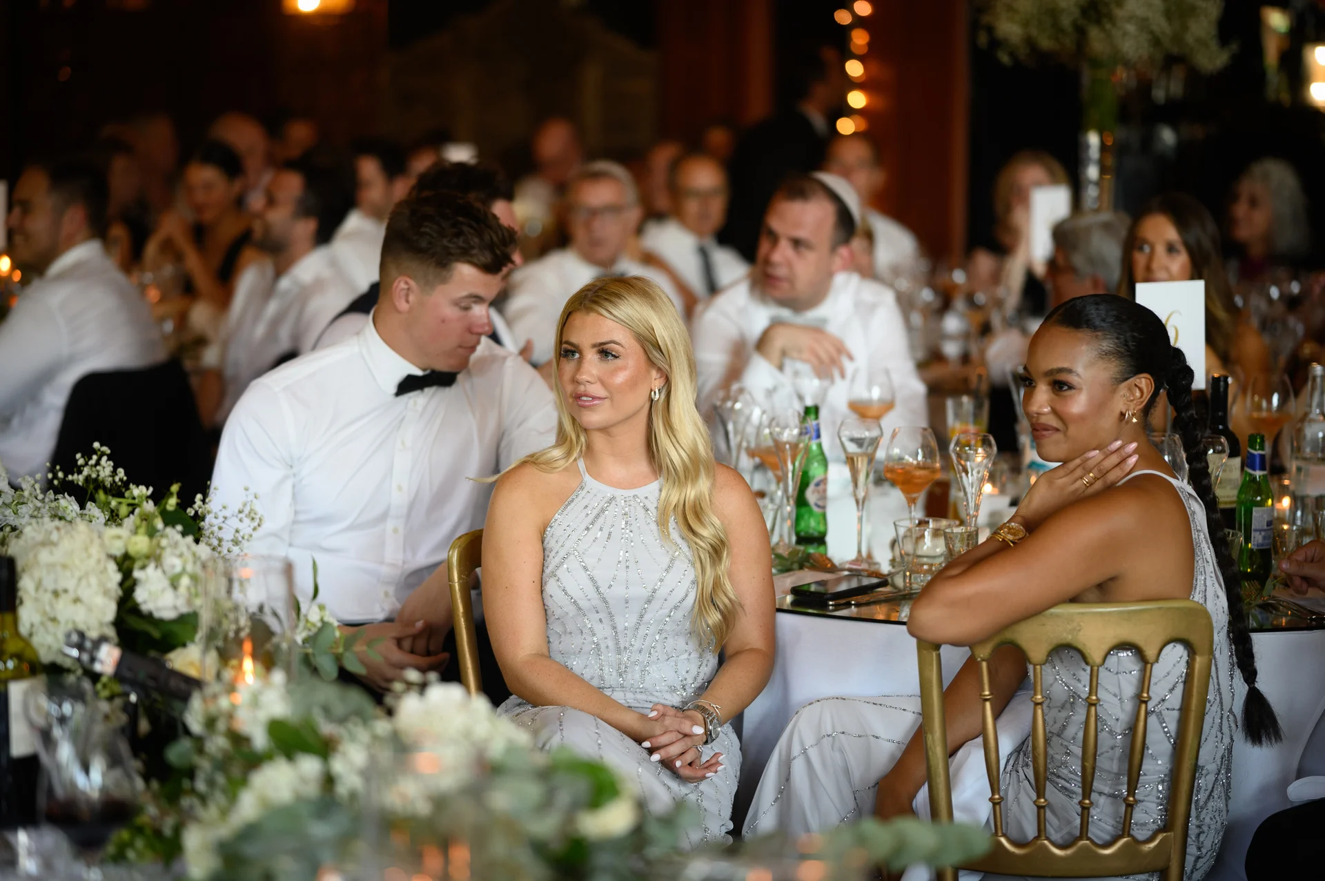 Guests are seated at beautifully decorated tables during a wedding reception, enjoying the meal amidst elegant floral centrepieces and warm ambient lighting.