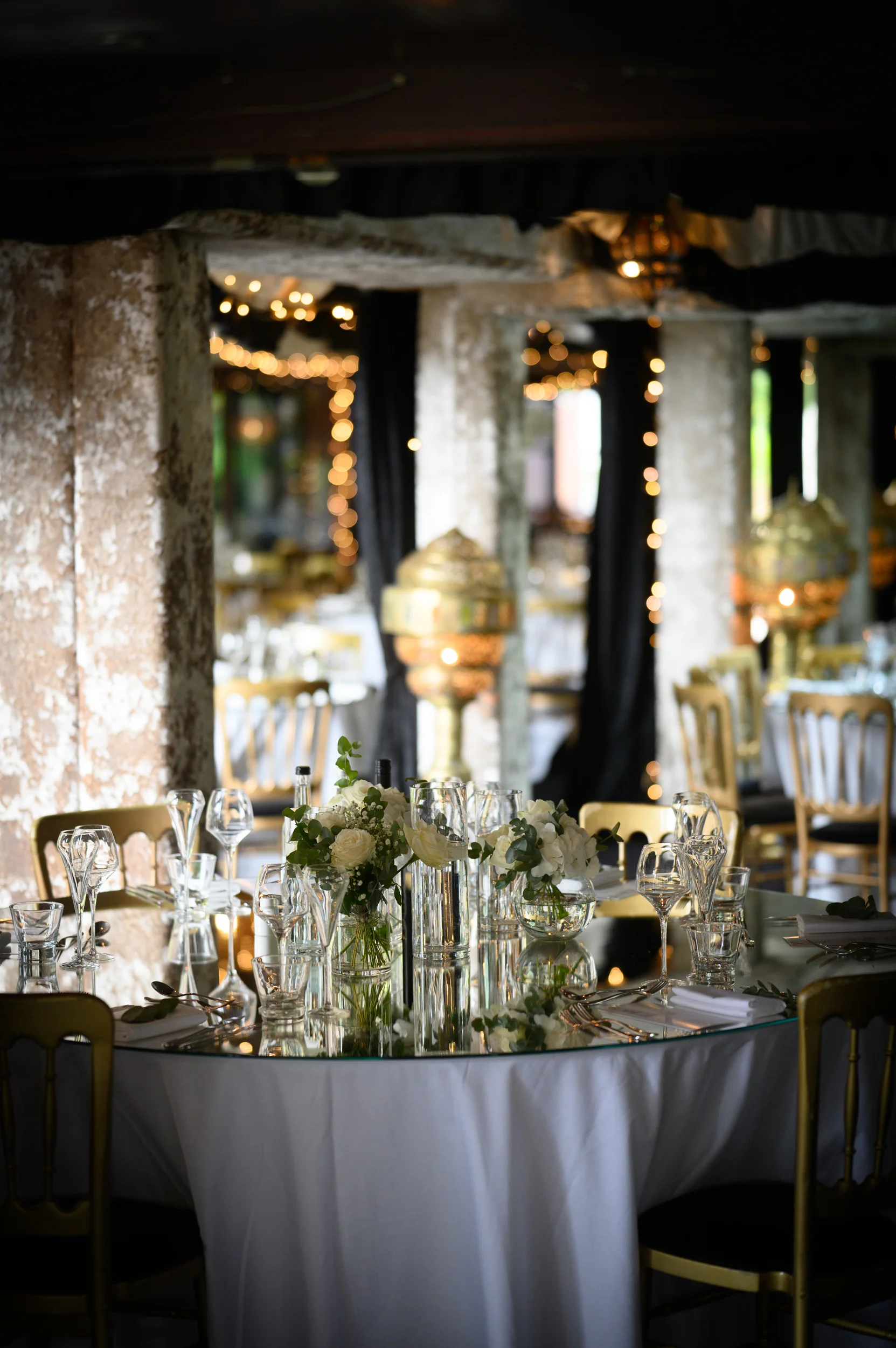 An elegantly styled round wedding table features a mirrored tabletop reflecting white rose floral centrepieces, delicate glassware, and soft candlelight.