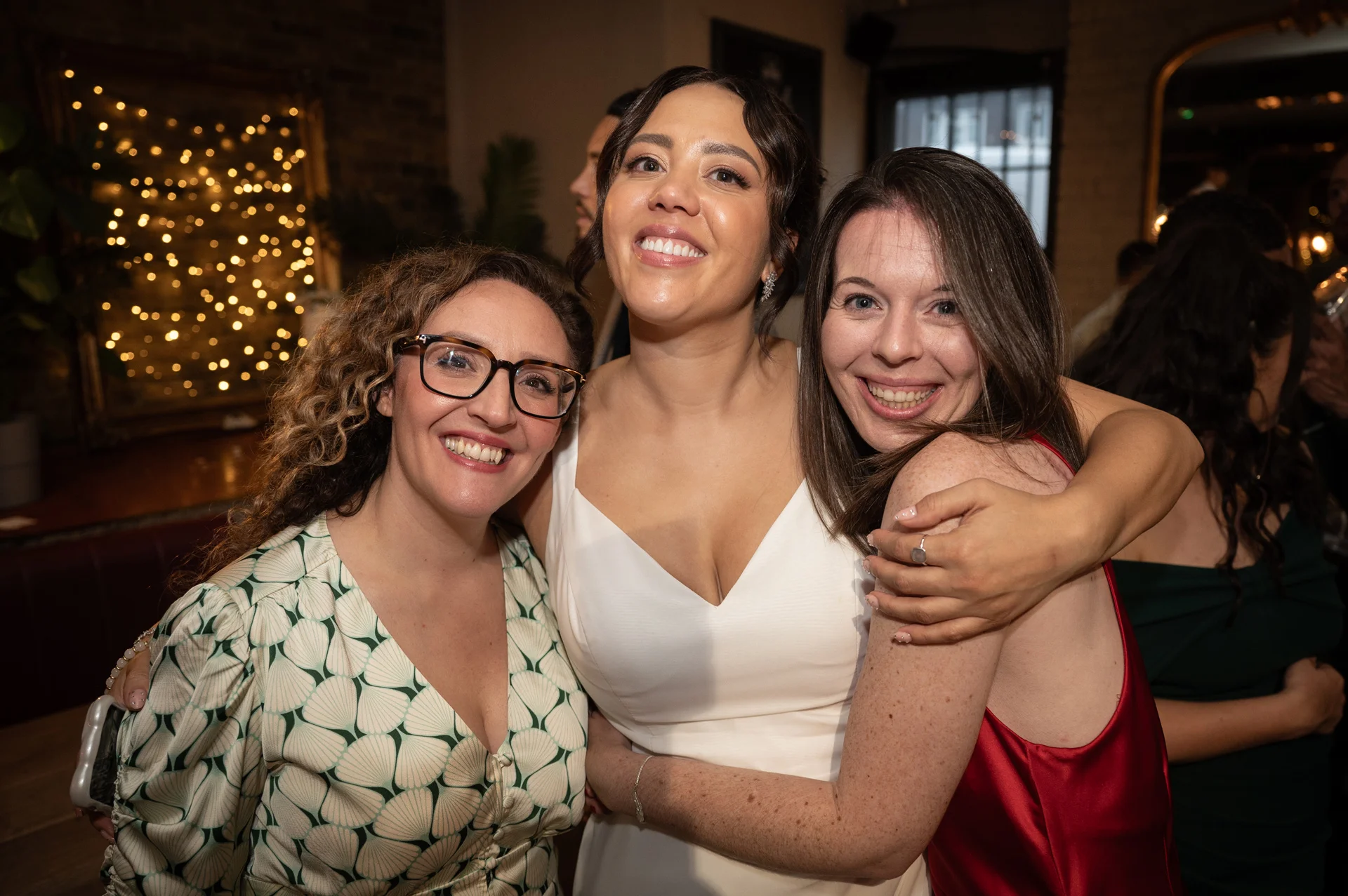 A joyful bride in a white gown embraces and laughs with two smiling guests at an evening wedding reception lit by direct flash.