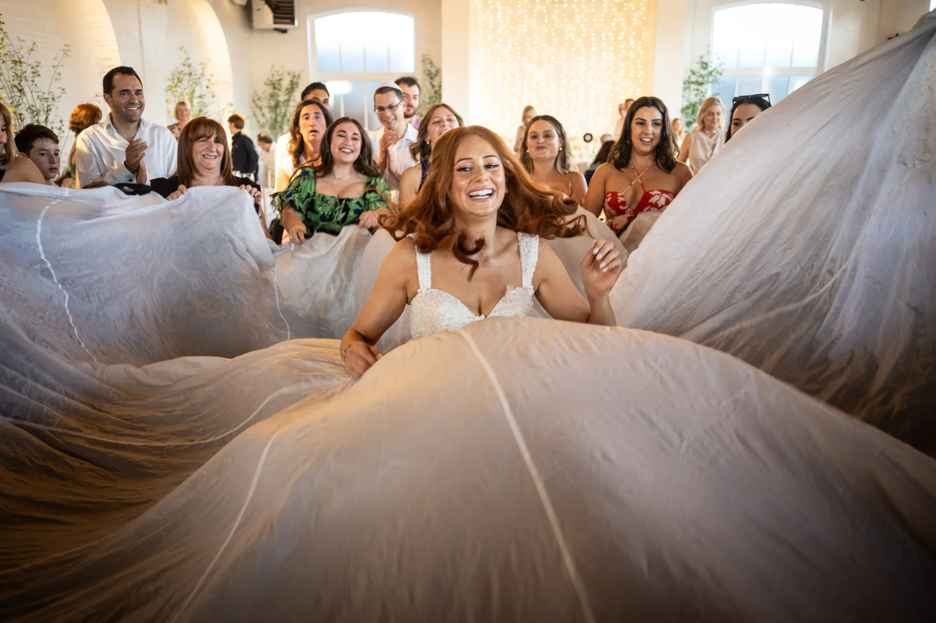 A joyful red-haired bride laughs while she and her wedding guests playfully toss a massive white veil and parachute-like fabric in a bright industrial venue.