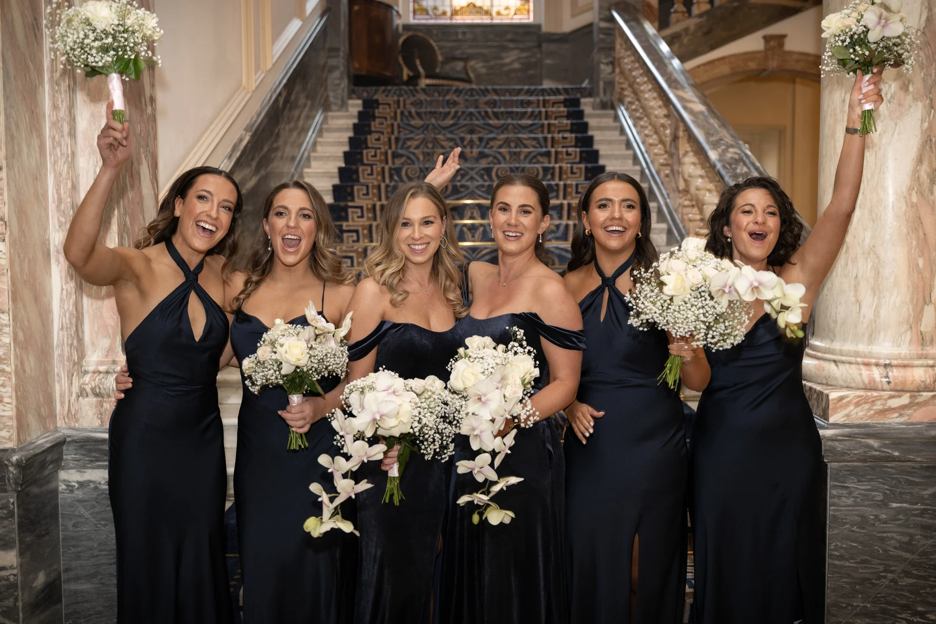 A joyful group of bridesmaids in matching navy blue dresses cheering and holding bouquets in front of a grand staircase inside a stately home.