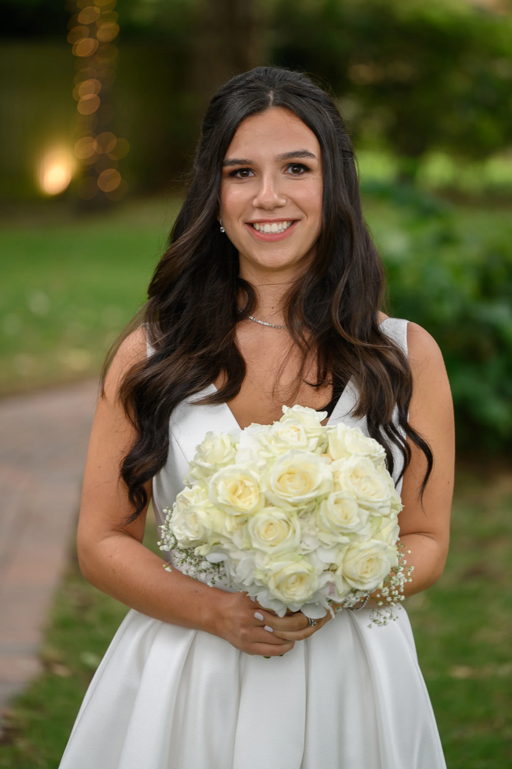 A smiling bride with dark hair styled in soft half-up waves holds a round bouquet of white roses and baby's breath outdoors.