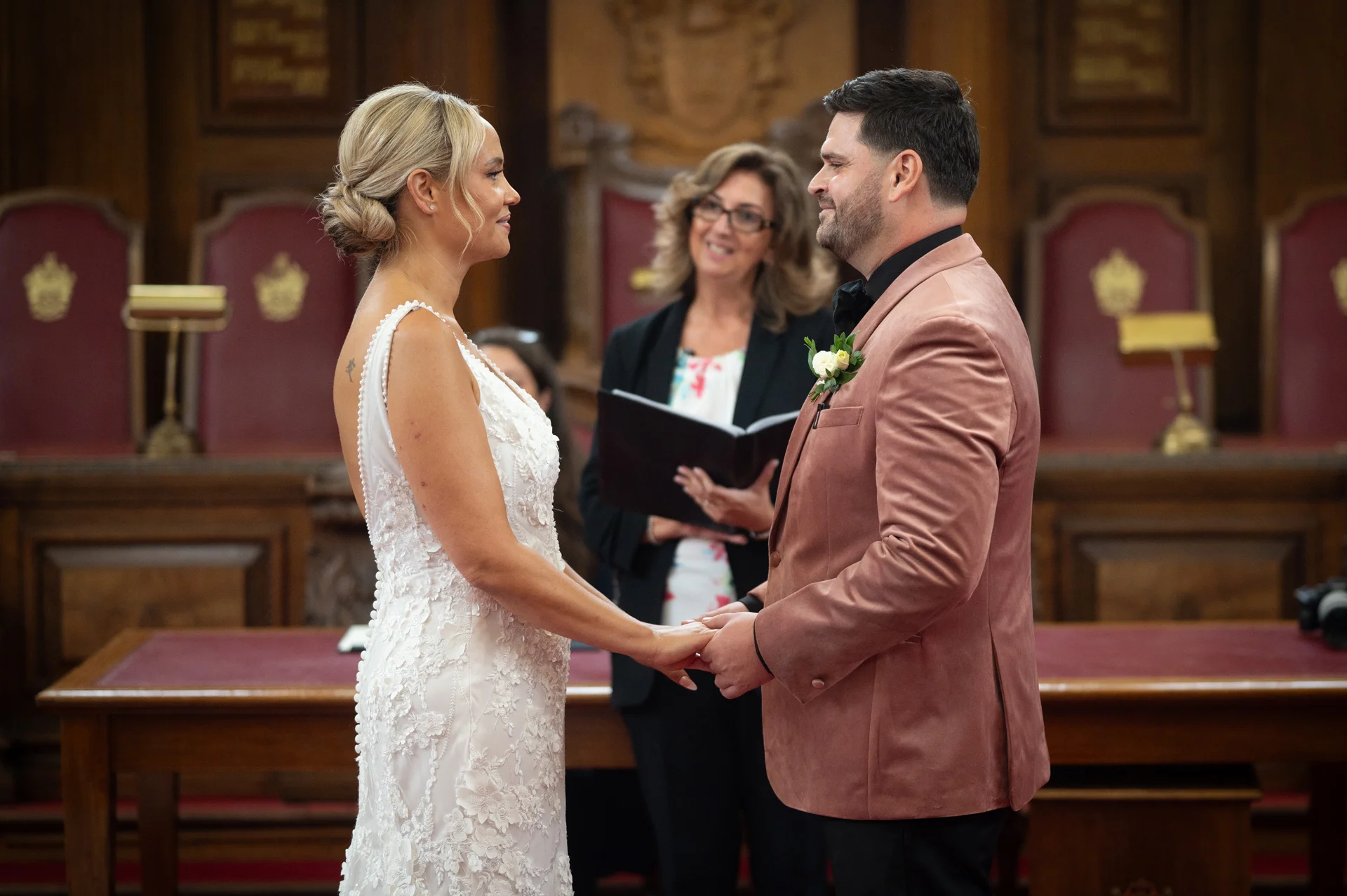 A bride in a floral appliqué gown and a groom in a pink velvet blazer hold hands and exchange vows during their civil ceremony inside a wood-panelled room.