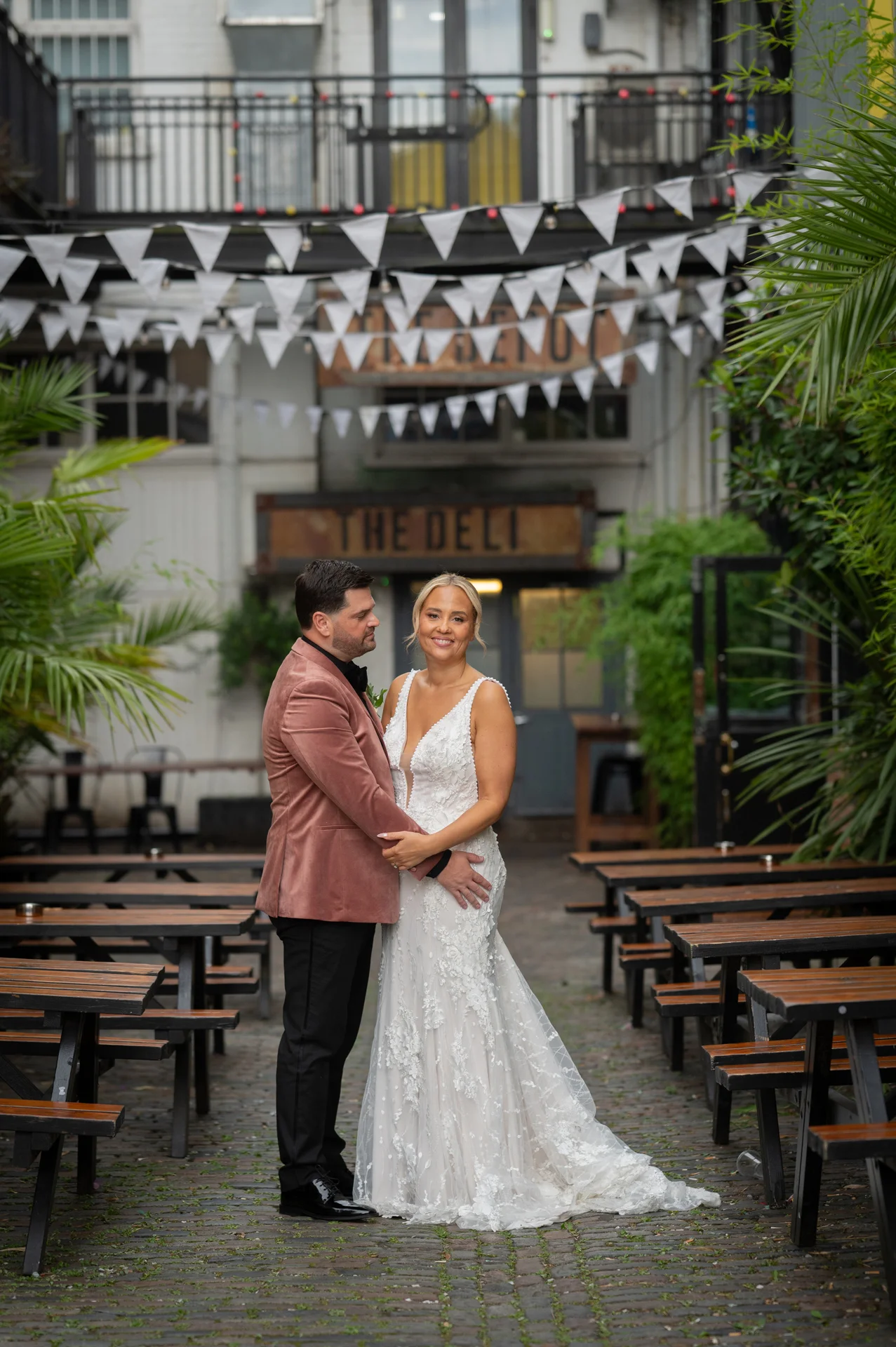 A bride and groom pose together in a cobbled urban courtyard decorated with bunting outside an industrial venue, with the bride wearing a floral applique gown and the groom in a pink velvet jacket.