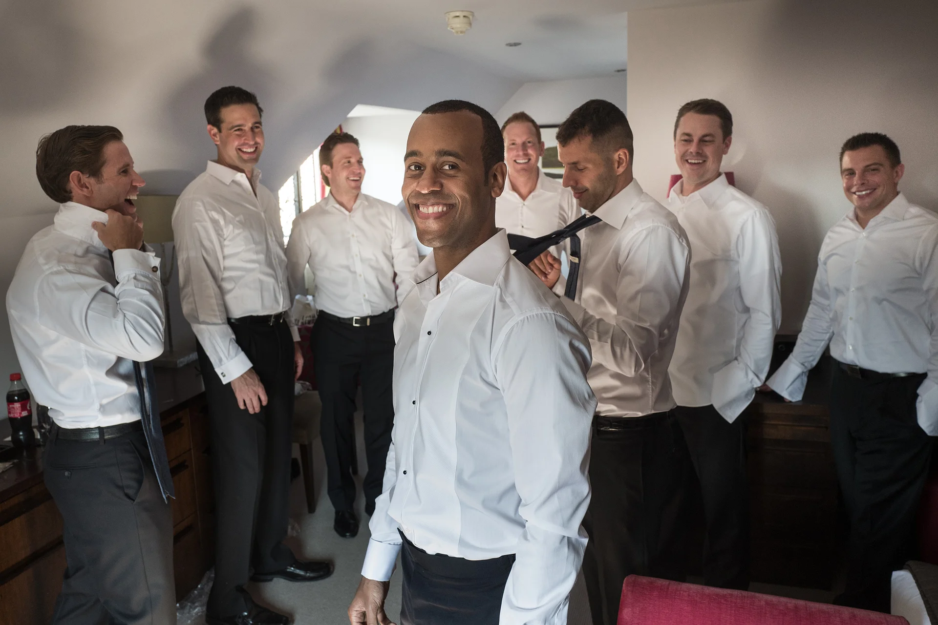 The groom stands smiling in the foreground wearing a white shirt while his groomsmen laugh and help each other get ready in a hotel room.