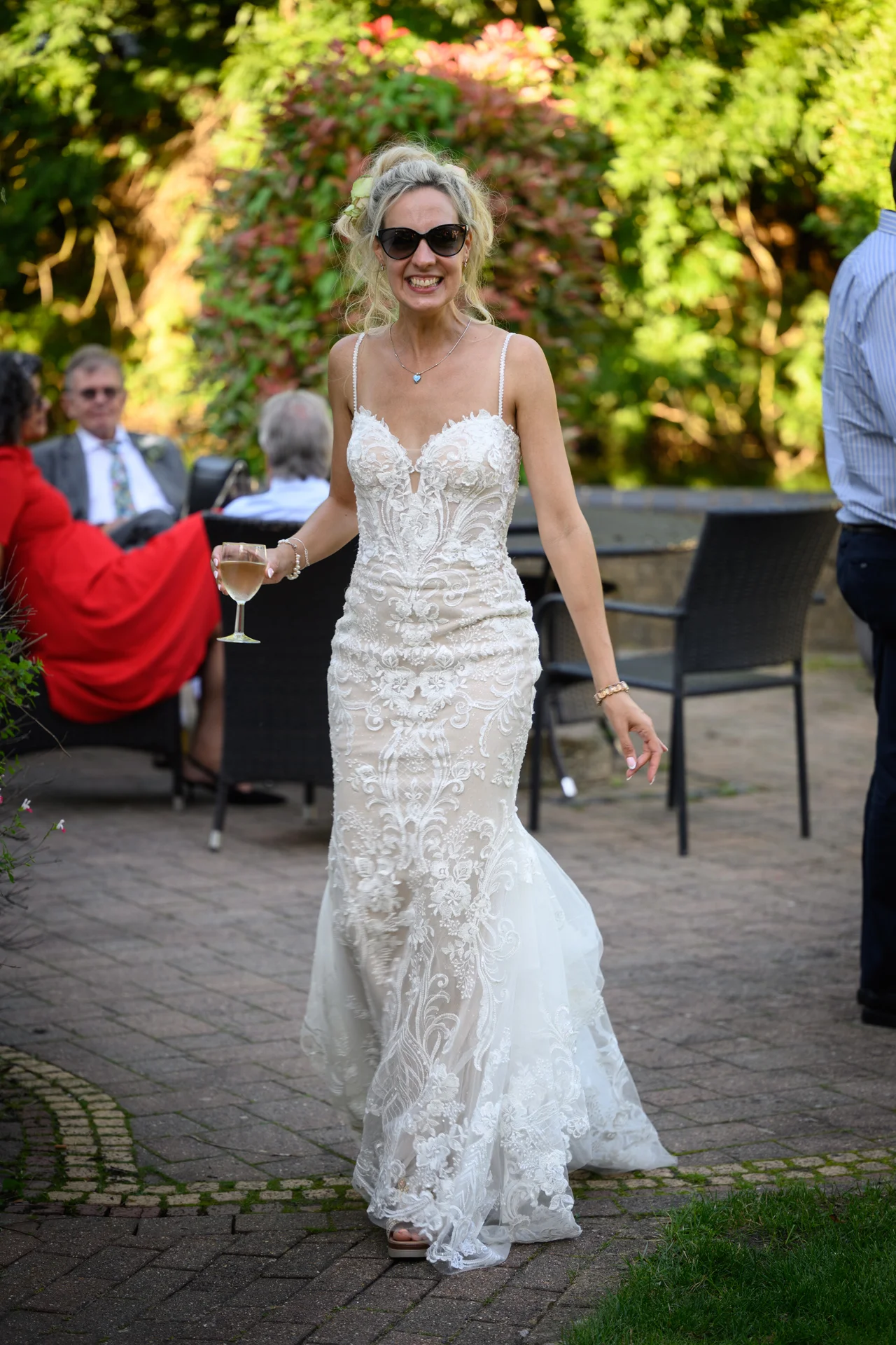 A joyful bride wearing sunglasses and a lace fit and flare wedding dress walks across a cobbled courtyard holding a glass of wine.