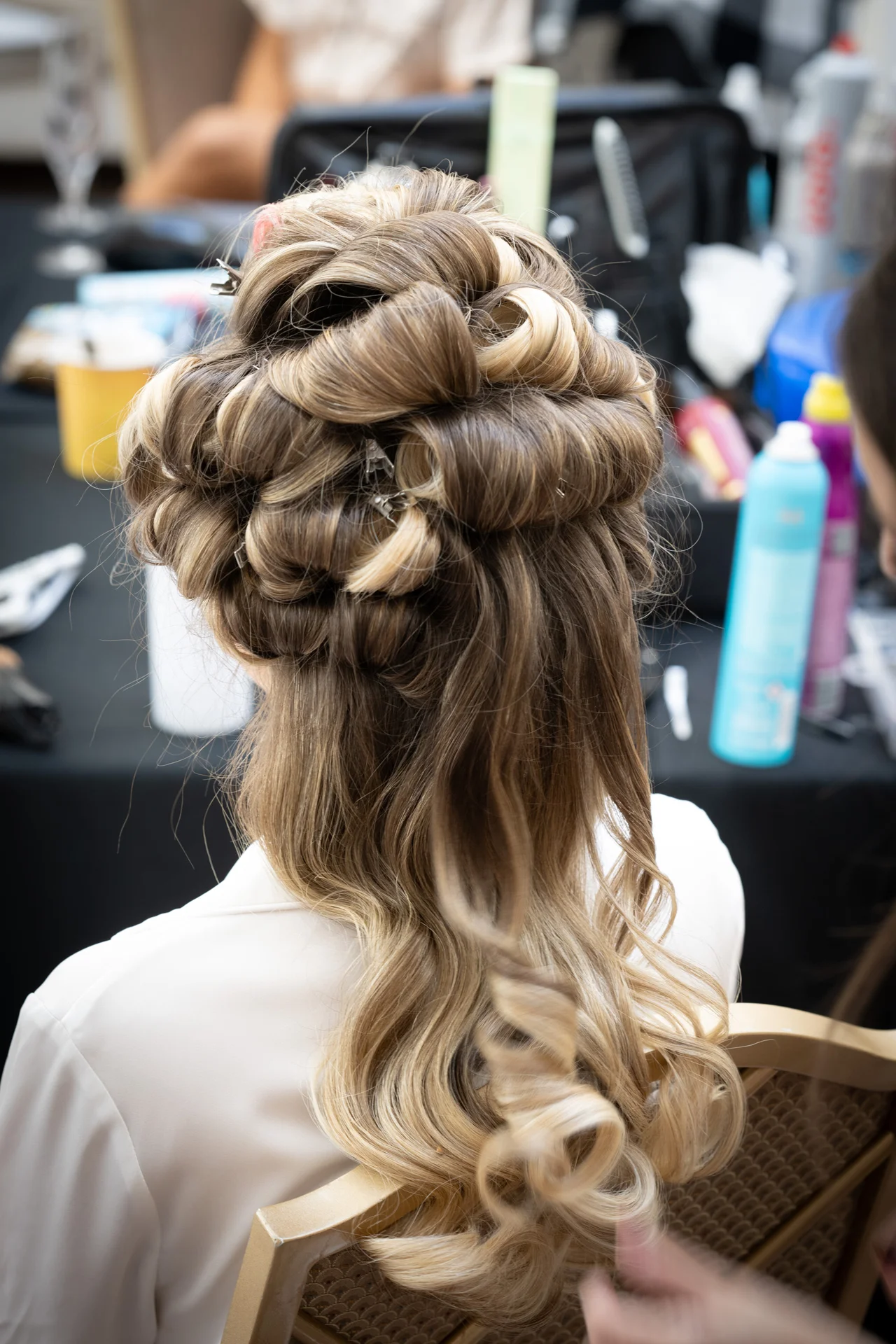 A close-up back view of a bride having her long blonde hair styled into an intricate half-up half-down look during morning wedding preparations.