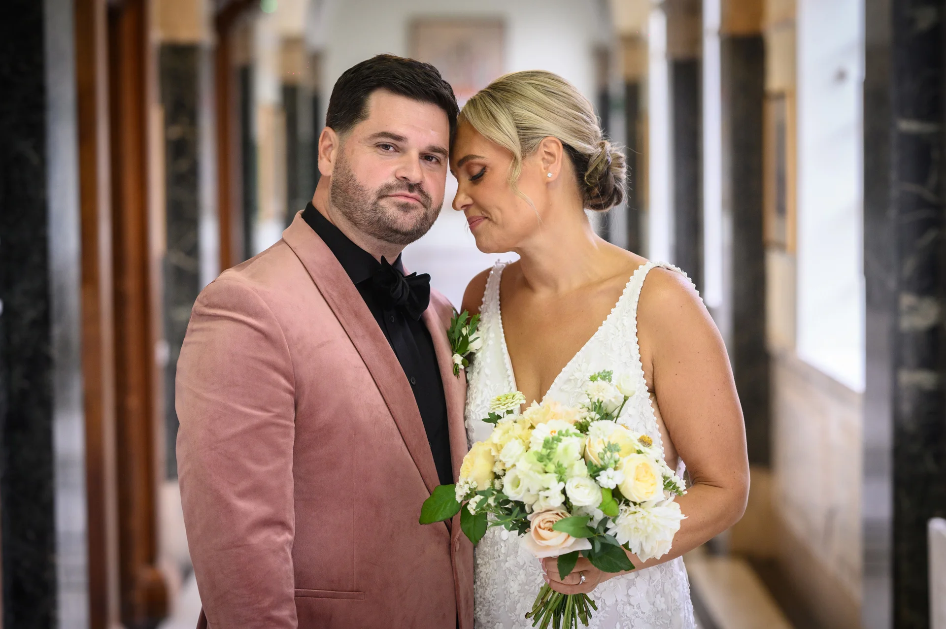 A groom wearing a pink velvet jacket and black shirt gently rests his head against the bride, who holds a cream and white floral bouquet in an elegant indoor corridor.
