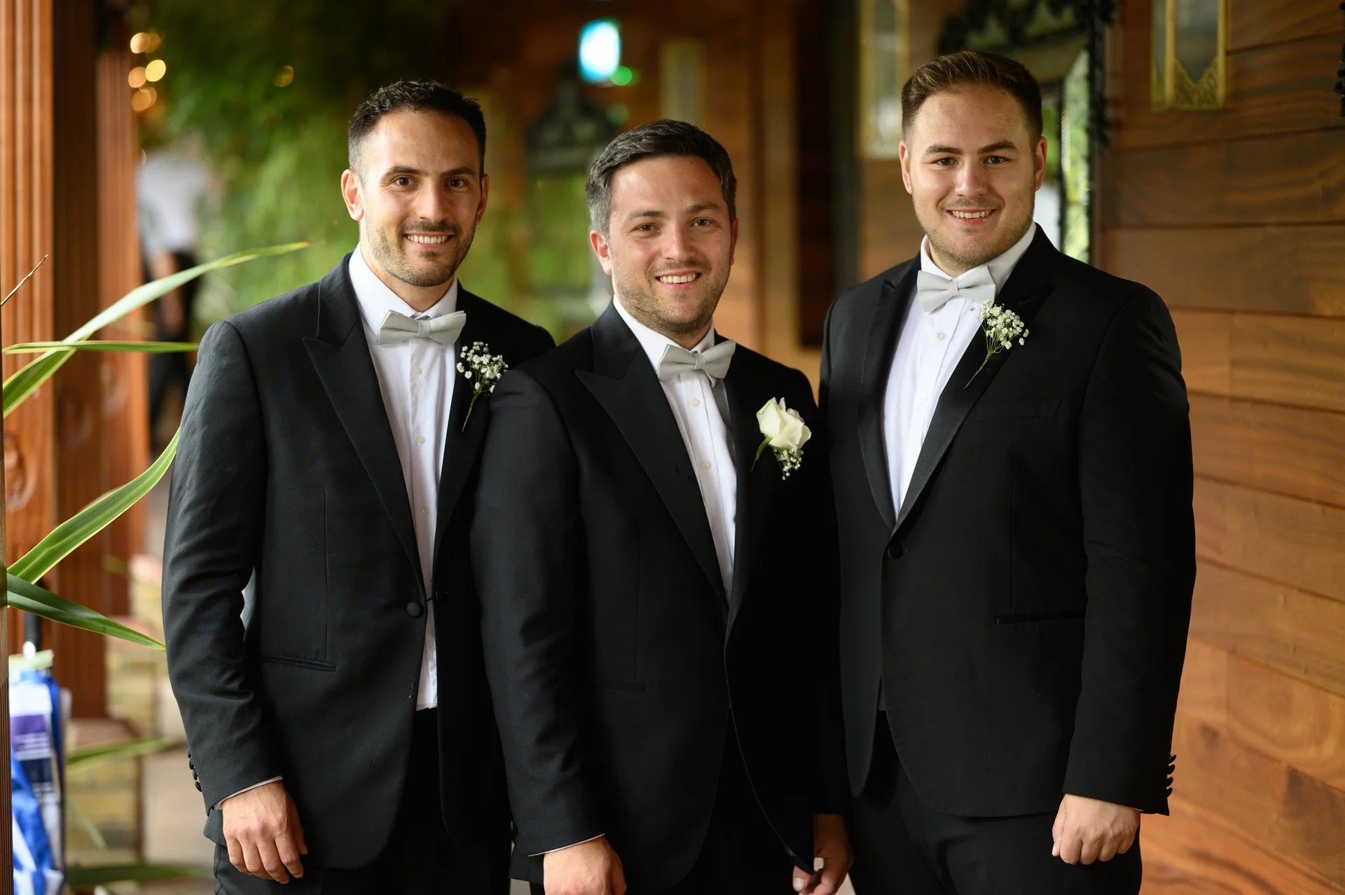 Three smiling groomsmen wearing classic black tuxedos with white shirts, pale bow ties and rose buttonholes pose outdoors against a wooden wall.