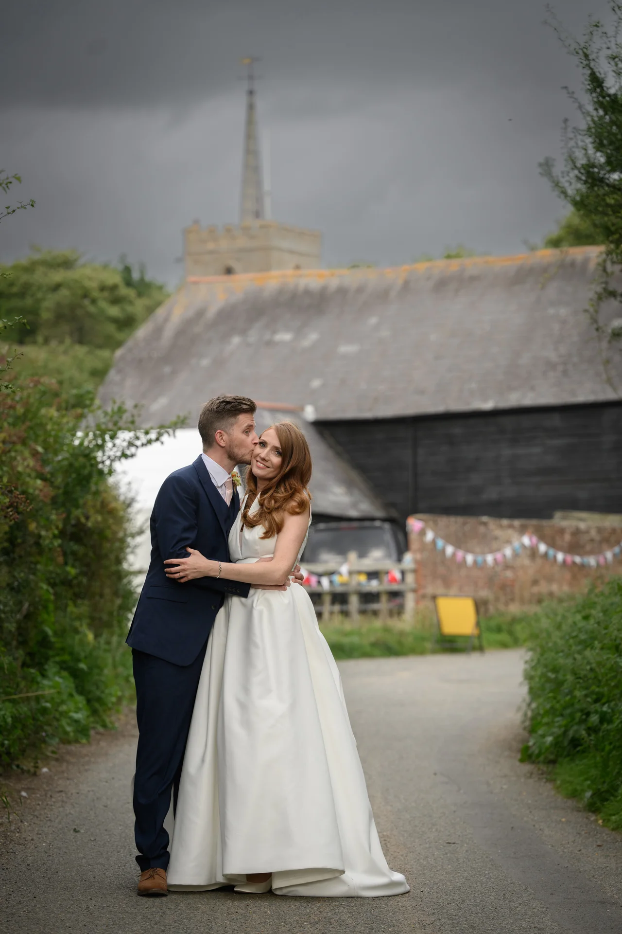 A groom kisses a smiling bride on the cheek outside a rustic barn and church under a dramatic overcast sky.