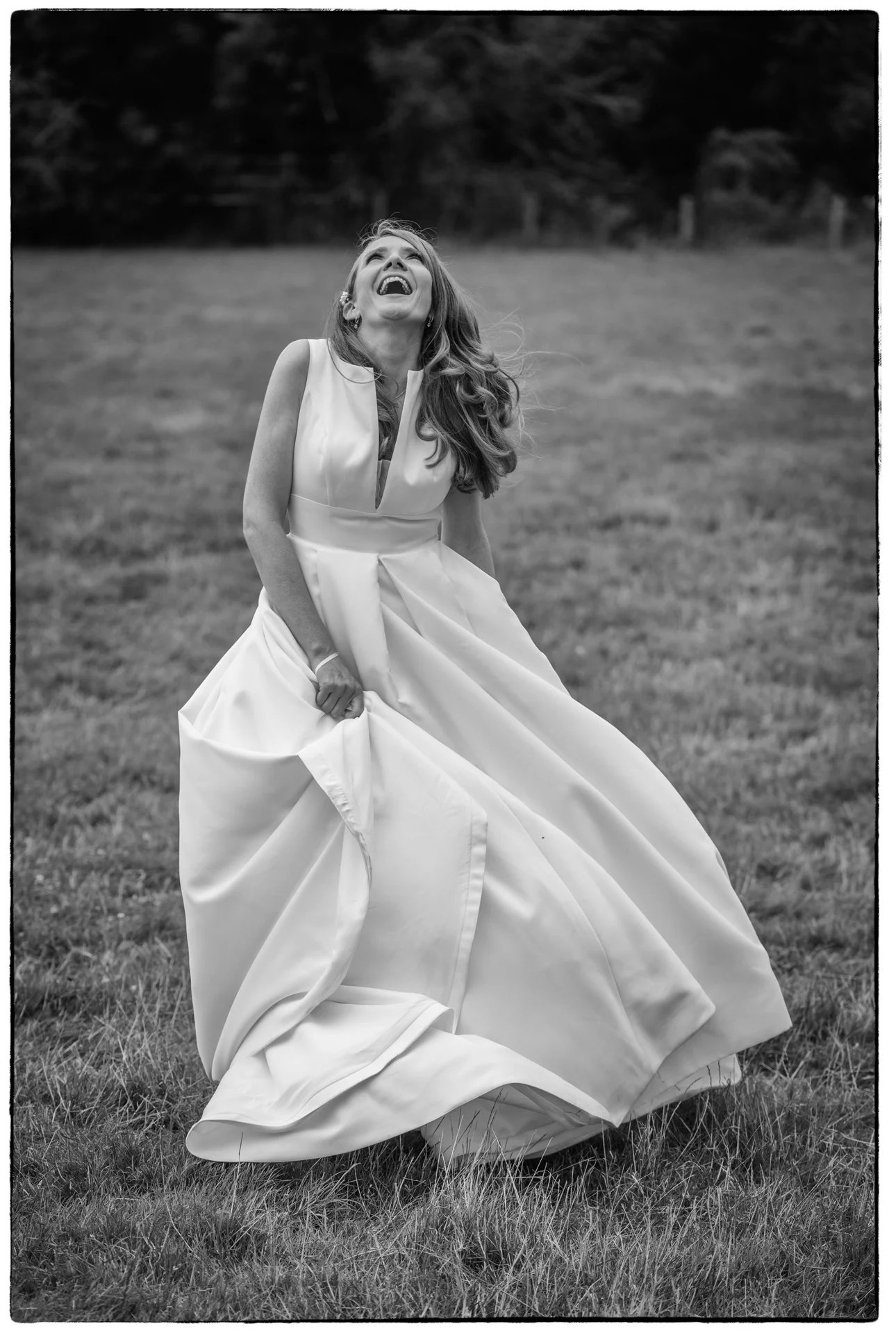 A joyous bride in a satin a-line gown laughs heartily while twirling in a grassy field in a black and white film style photograph.