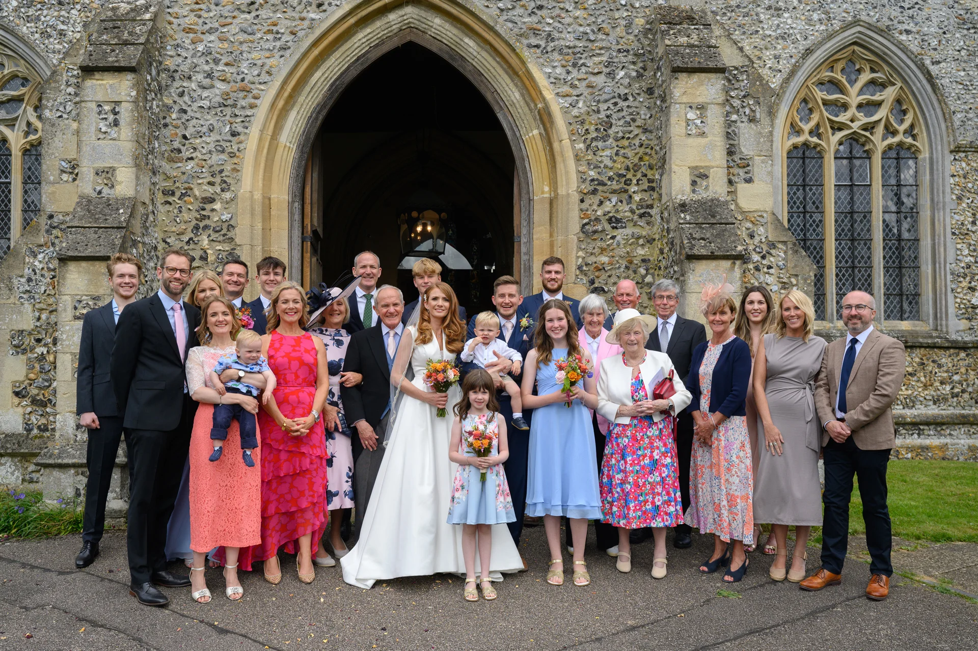 A large wedding party and family group pose smiling outside the historic stone entrance of an English country church.