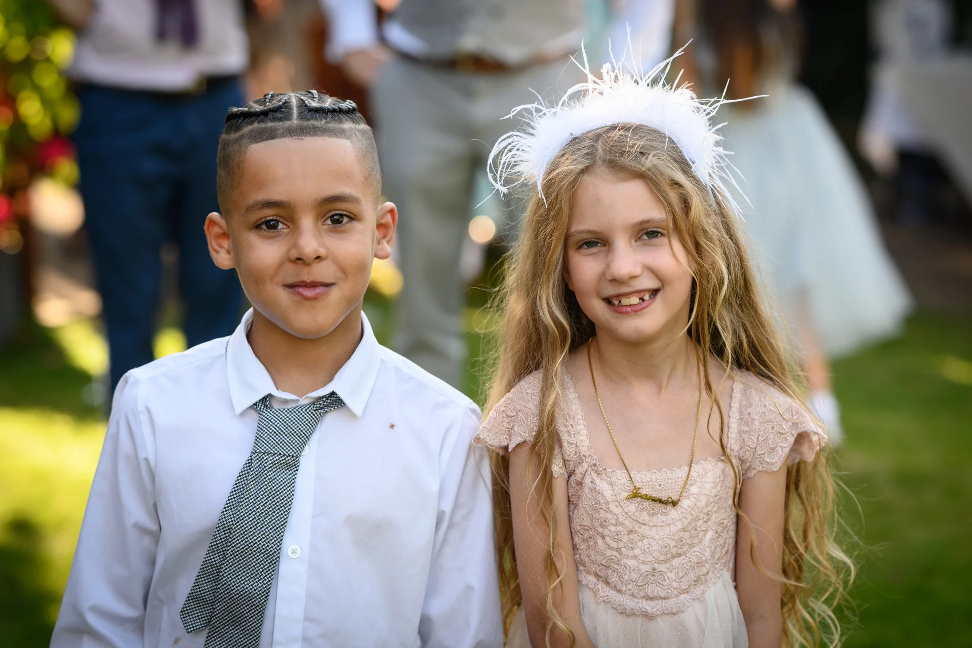 A young page boy and flower girl smile happily for the camera outdoors in natural light during a wedding reception.