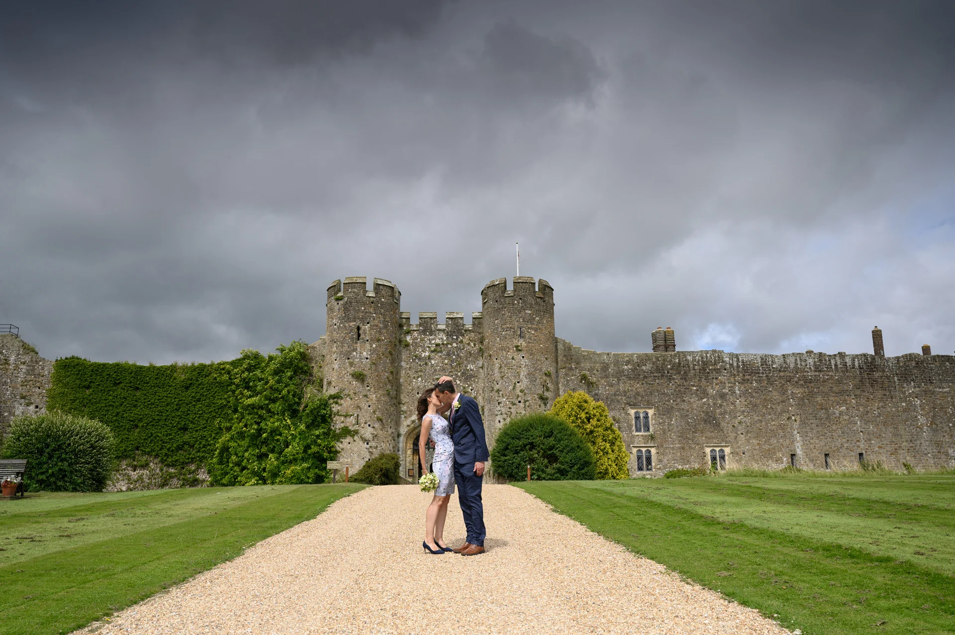 A newlywed couple kisses on a gravel path leading to a grand stone castle under a dramatic, cloudy sky.