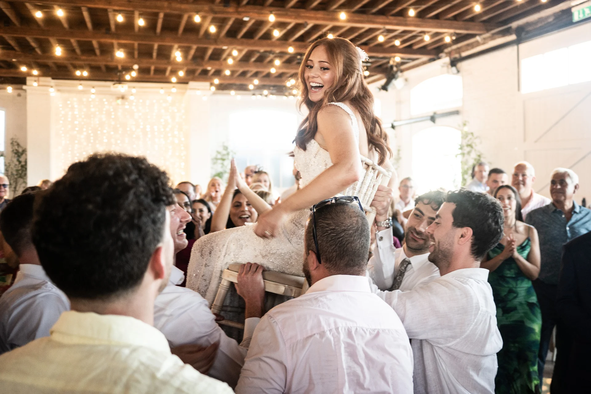 A joyful bride is lifted high on a chair during the wedding reception celebration inside a bright warehouse venue with exposed rafters.