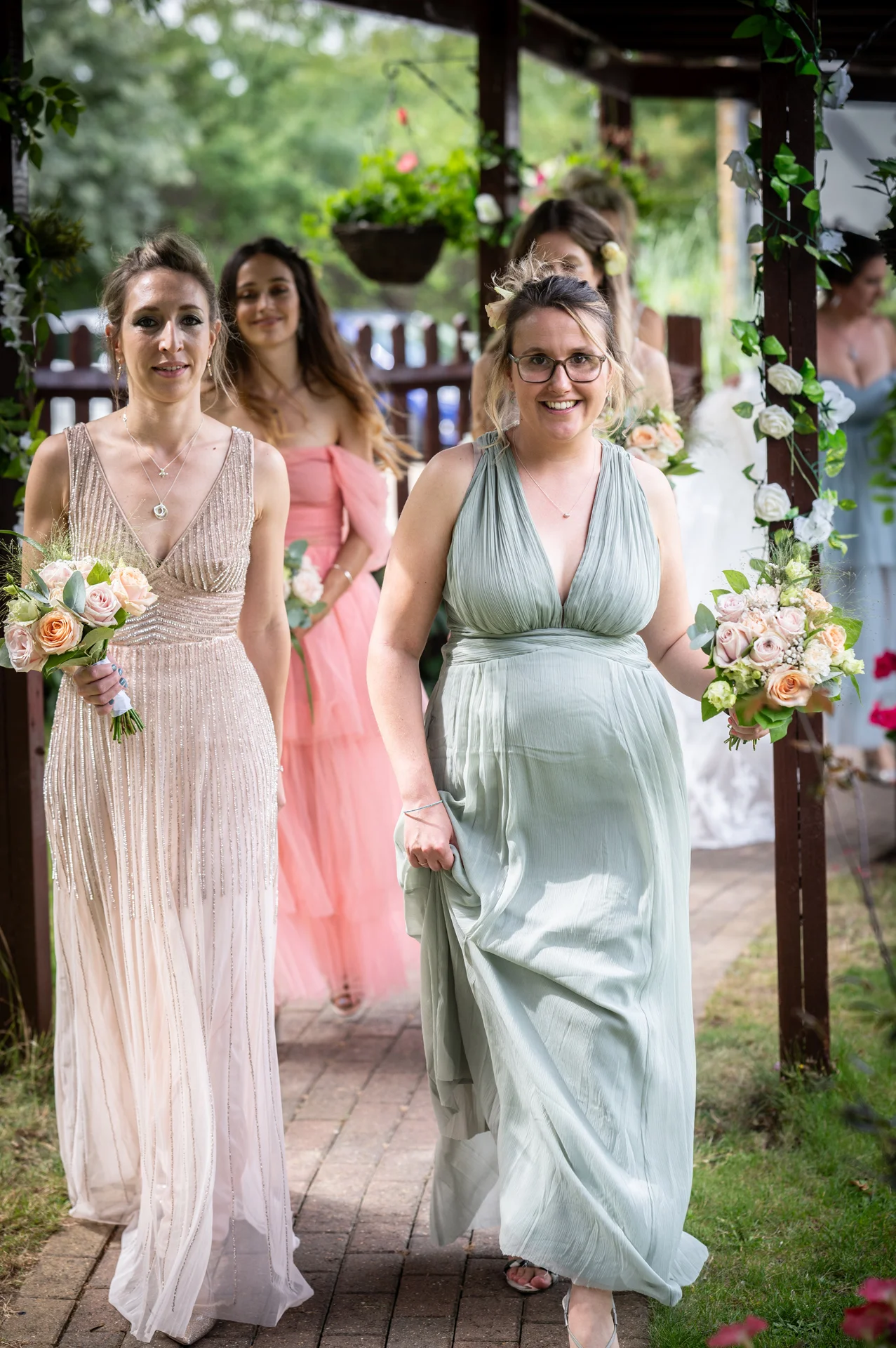 A group of smiling bridesmaids in mismatched dresses carrying floral bouquets walk along an outdoor brick pathway.