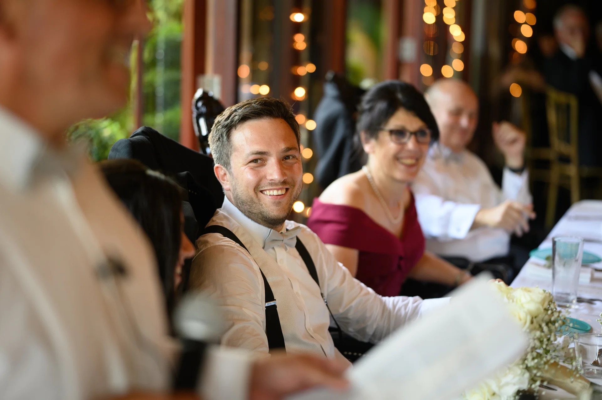 A groom in a white shirt and braces smiles happily at the wedding reception while listening to speeches under a fairy light canopy.