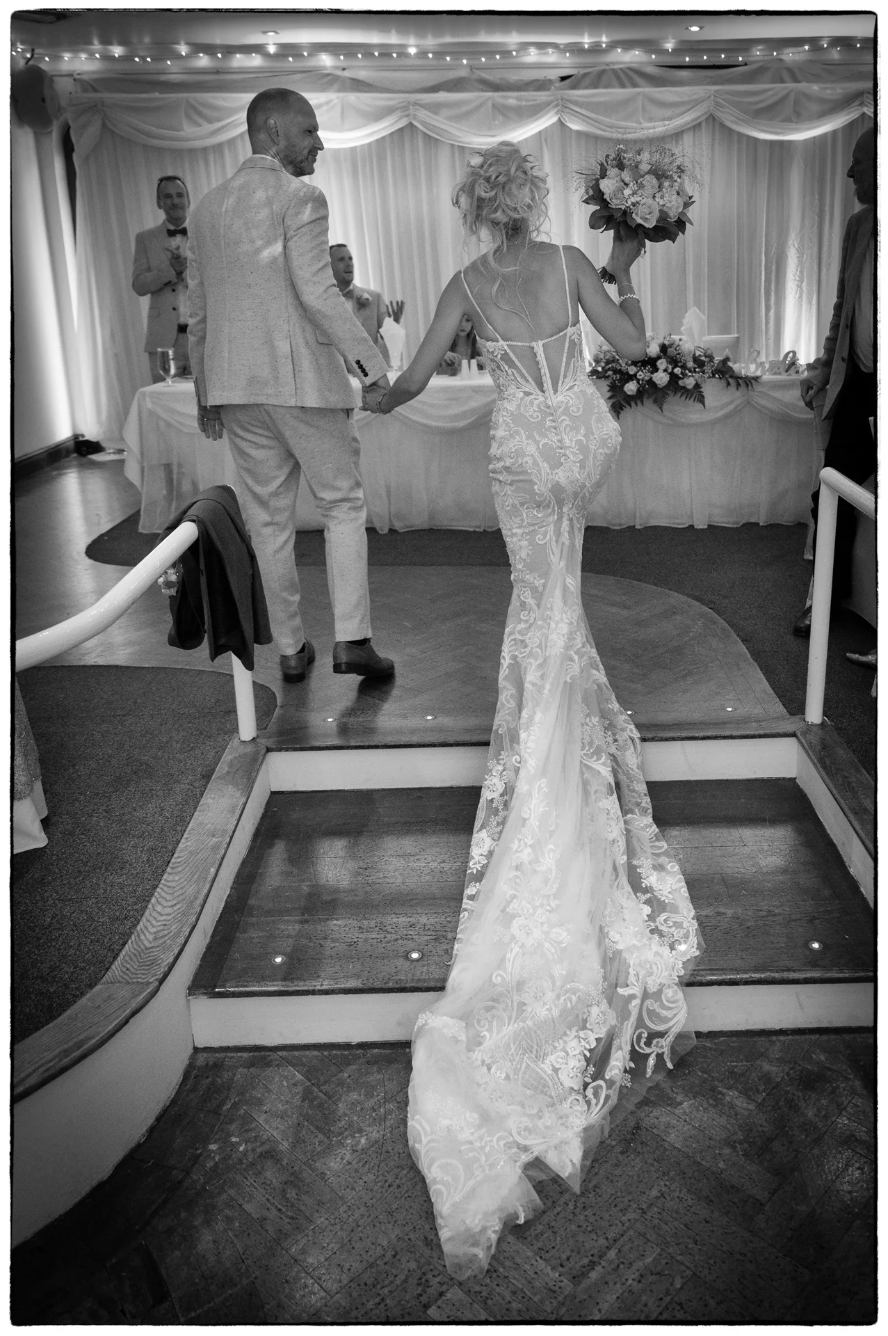 The newlywed couple walks hand in hand down a wooden staircase in a reception venue, with the bride wearing a fitted lace gown with a long train and carrying a bouquet.